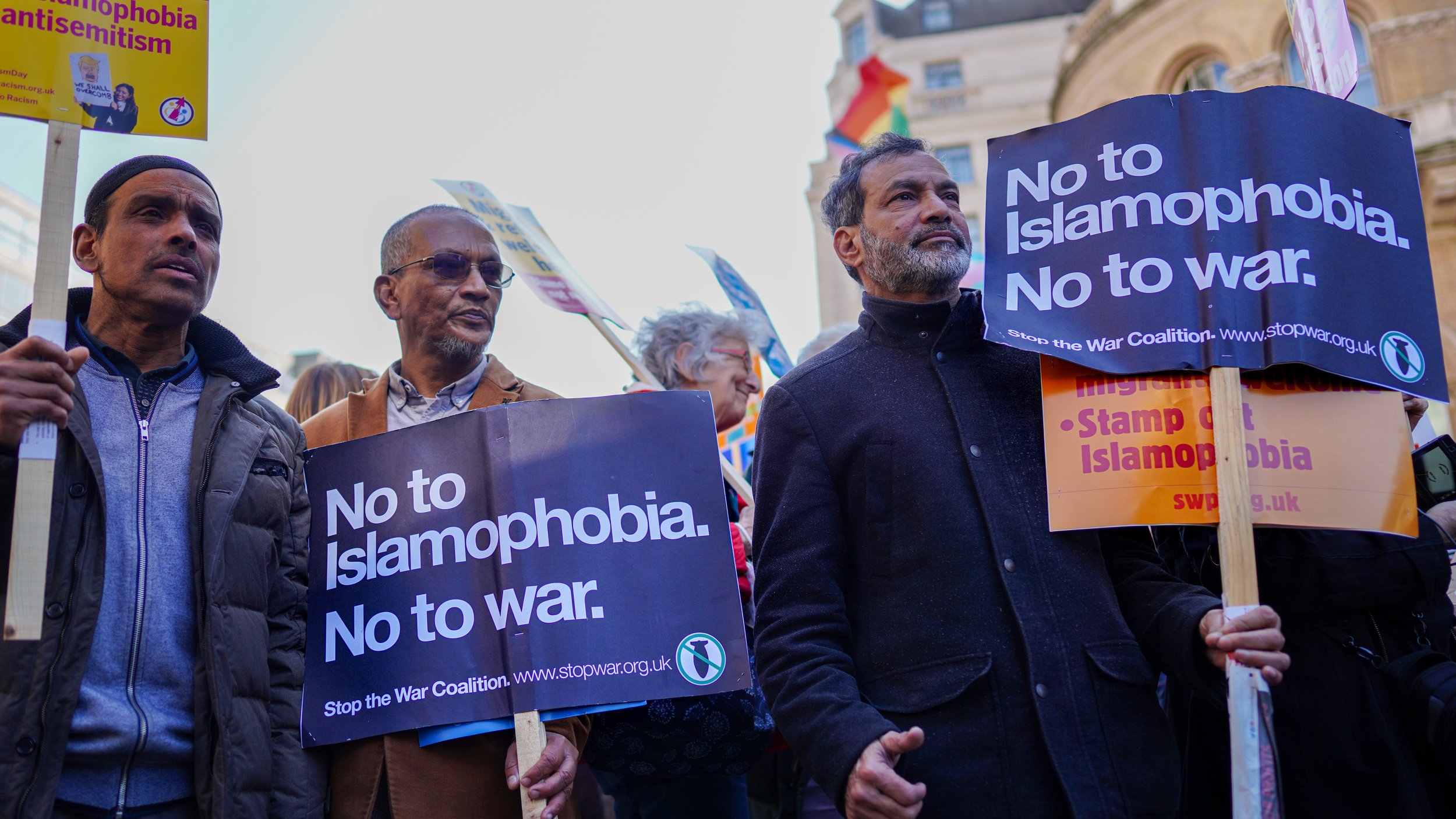 L'immagine mostra un gruppo di persone che partecipano a una manifestazione. Alcuni di loro stanno tenendo cartelli che riportano messaggi come "No to Islamophobia. No to war." e "Stop the War Coalition." L'atmosfera è di protesta pacifica, con i manifestanti che esprimono i loro punti di vista contro l'islamofobia e le guerre. In lontananza, si possono notare delle bandiere, che aggiungono un senso di diversità e unità all'evento.