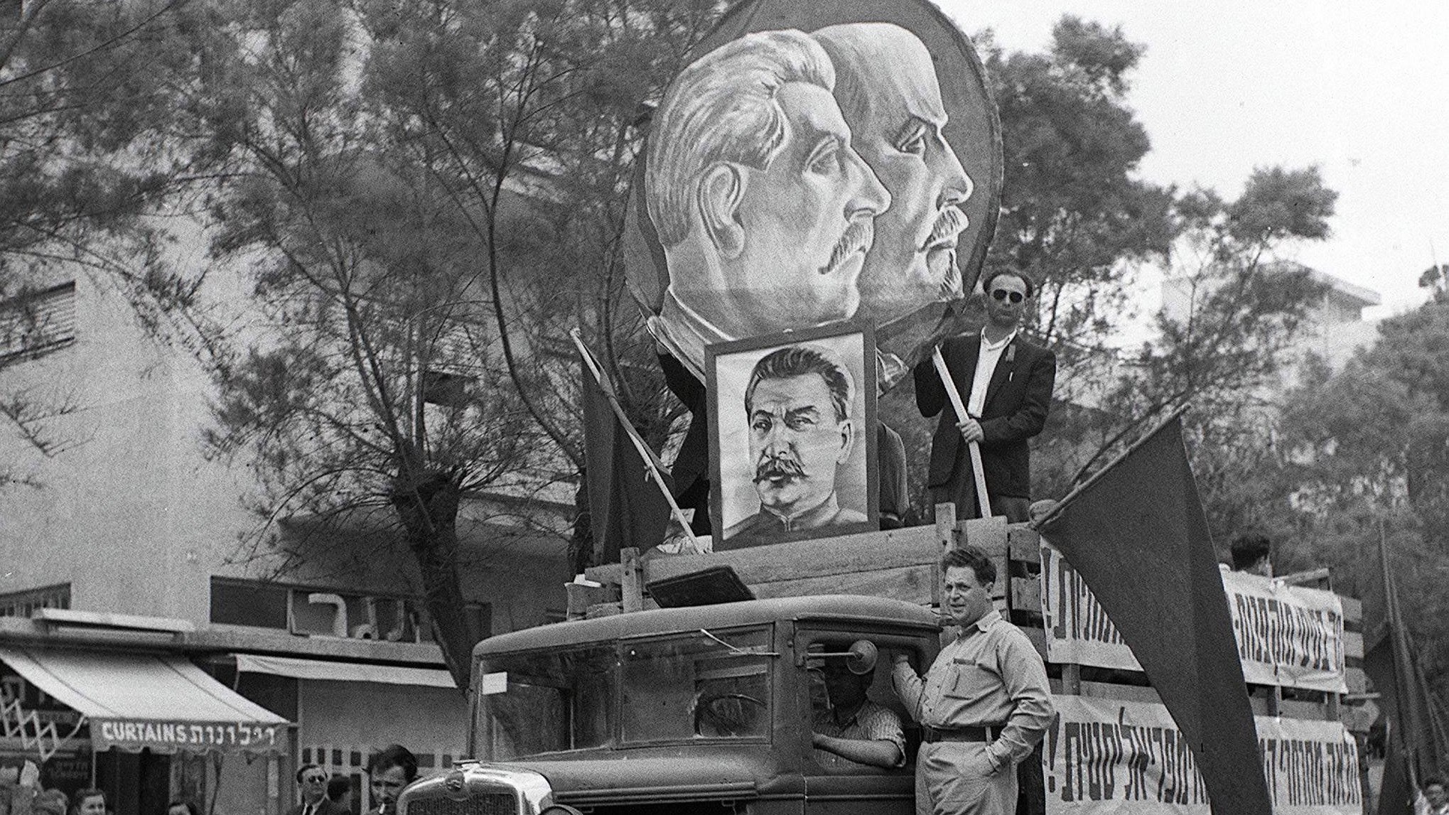 تصویر نشاندهنده یک خودروی قدیمی است که در یک مراسم راهپیمایی یا تظاهرات شرکت کرده است. بر روی ماشین بزرگنمایی از چهرههای افرادی با موهای کوتاه و مقدار زیادی نماد و پرچمهایی وجود دارد. فردی در حالت ایستاده بر روی خودرو قرار دارد و به نظر میرسد که در حال صحبت یا اشاره به تماشاگران است. پسزمینه شامل گروهی از مردم است که در کنار خیابان ایستادهاند، و فضایی از شور و اشتیاق را در این رویداد به تصویر میکشد.