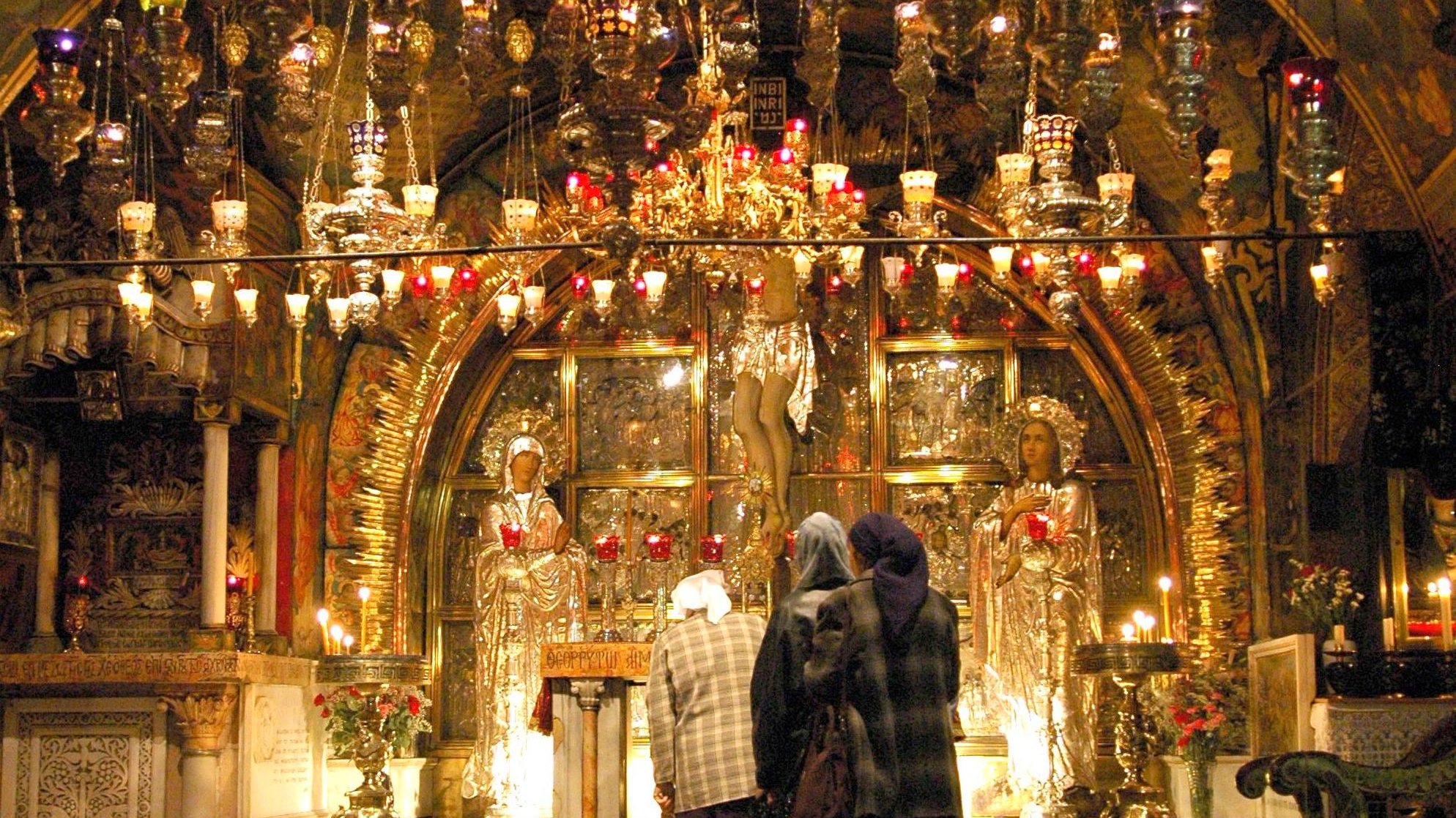 L'image montre l'intérieur d'un lieu de culte, probablement une église ou une chapelle. L'architecture est ornée, avec un plafond voûté richement décoré. On peut voir des lumières colorées suspendues et des candélabres, créant une ambiance chaleureuse et mystique. Au centre, il y a des figures dorées, possiblement des icônes ou des statues, entourées de bougies et de fleurs. Quelques personnes, vêtues de manteaux et de foulards, se tiennent devant cette scène, semblant s'engager dans une prière ou une contemplation.