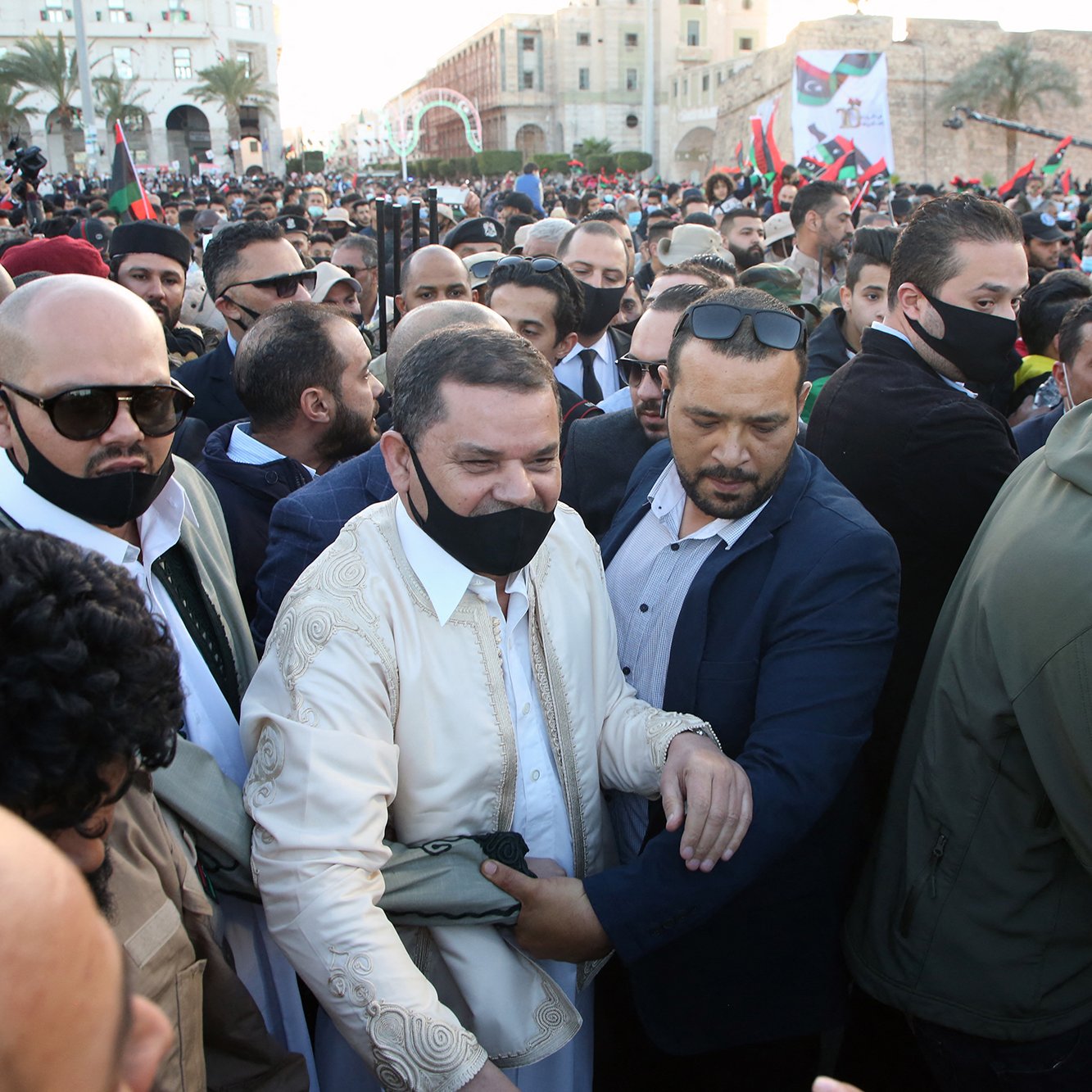 الصورة تُظهر حشودًا كبيرة من الناس في مكان عام، بعضهم يرتدي الكمامات، ويبدو أن هناك شخصيات بارزة في الوسط. يظهر الأشخاص في الصورة ممالأة وتفاعل مع بعضهم، مما يوحي بأن هناك حدثًا مهمًا يجري. يمكن أن يكون الحدث مظاهرة أو تجمعًا سياسيًا، حيث تحمل الأعلام وتظهر بعض الوجوه المألوفة. الجو بشكل عام يبدو متوترًا ولكن مليئًا بالحماس.