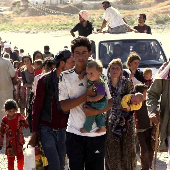 L'image montre un grand groupe de personnes se déplaçant, probablement dans un contexte de crise. On voit des adultes et des enfants marchant, avec des expressions sérieuses sur leurs visages. Certains portent des objets, et il y a une camionnette dans le fond. L'environnement semble désolé, avec peu d'infrastructures visibles. Cette scène évoque des thèmes de déplacement et de lutte, typiques de situations de conflit ou de catastrophe.