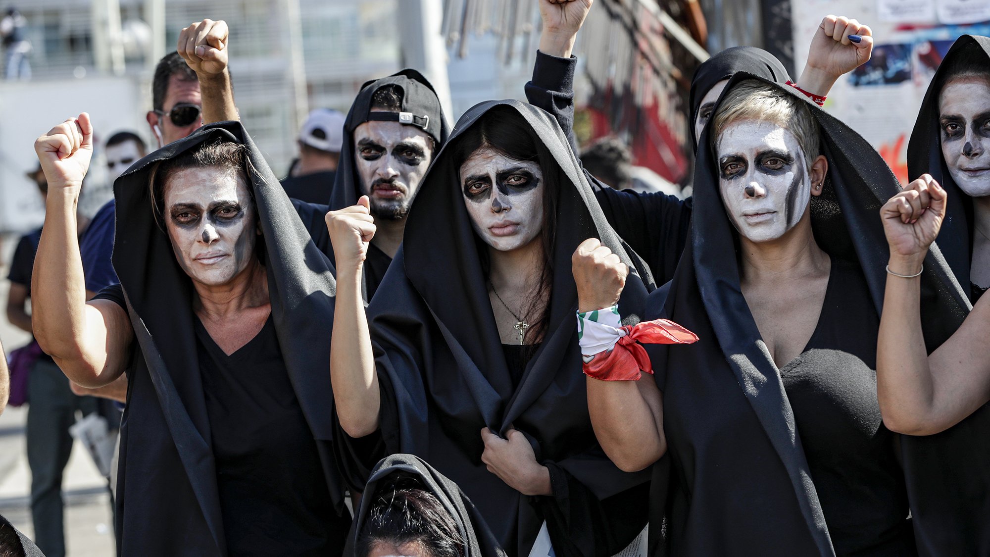 در این تصویر، گروهی از افراد زن و مرد را مشاهده می‌کنیم که لباس‌های سیاهی به تن دارند و صورت‌های خود را با رنگ سفید نقاشی کرده‌اند. آن‌ها در حال نمایش اعتراض خود هستند و برخی از آن‌ها با دستان بلند شده، نشانه‌ای از قدرت و اتحاد را نشان می‌دهند. پس‌زمینه تصویر به وضوح حاکی از یک تجمع یا تظاهرات است. حالت چهره‌ها جدی و معنادار به نظر می‌رسد، که بیانگر عزم و اراده آن‌ها برای بیان دیدگاه‌ها و خواسته‌هایشان است.