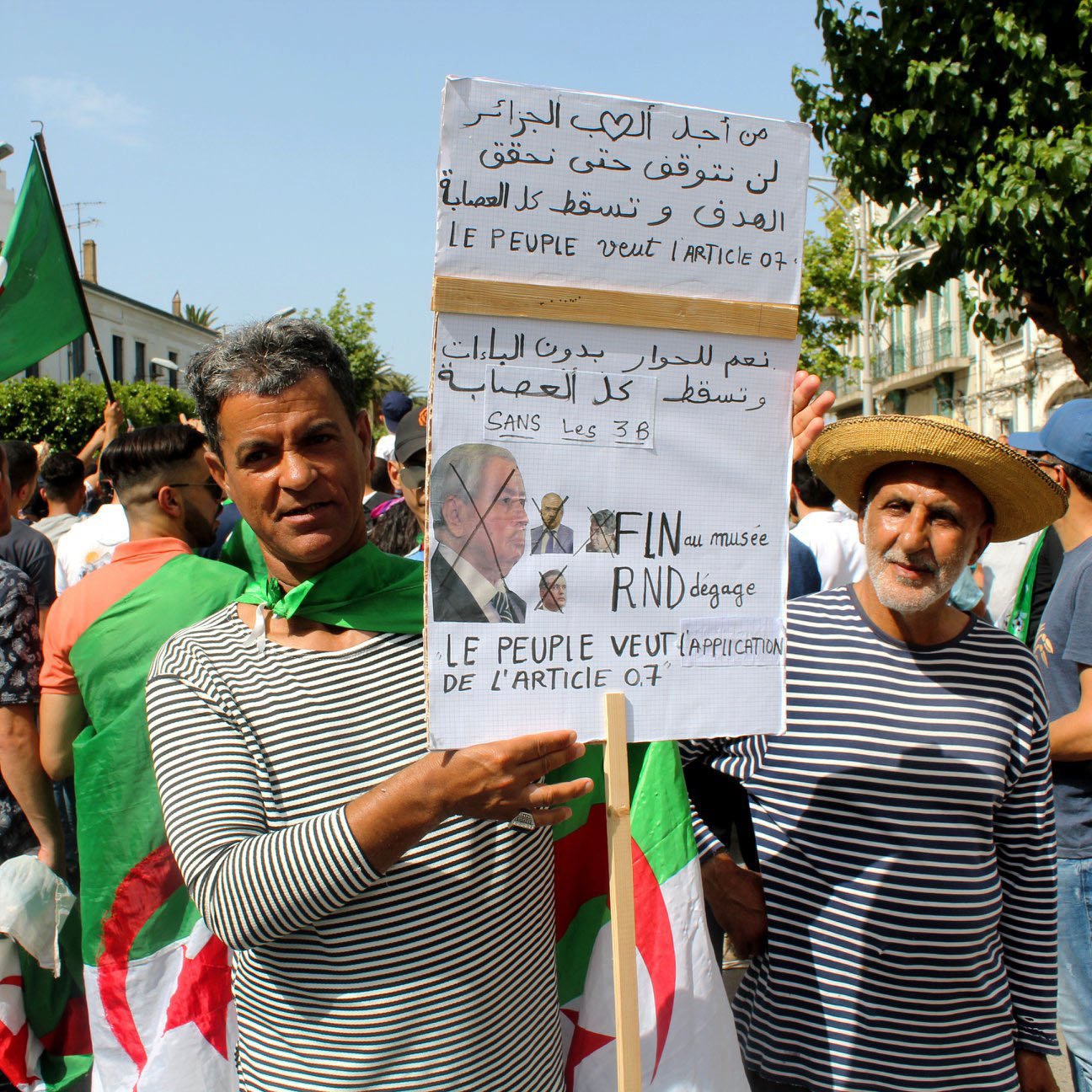 L'image montre un groupe de personnes manifestant, principalement des hommes, certains portant des drapeaux algériens. Deux manifestants se tiennent en avant-plan, l'un tenant une pancarte avec des inscriptions en arabe et en français. La pancarte semble exprimer des revendications politiques, notamment la fin de certaines pratiques liées à la politique algérienne. Les manifestants semblent unis et déterminés, se rassemblant pour faire entendre leur voix. L'atmosphère générale est celle d'une protestation pacifique.