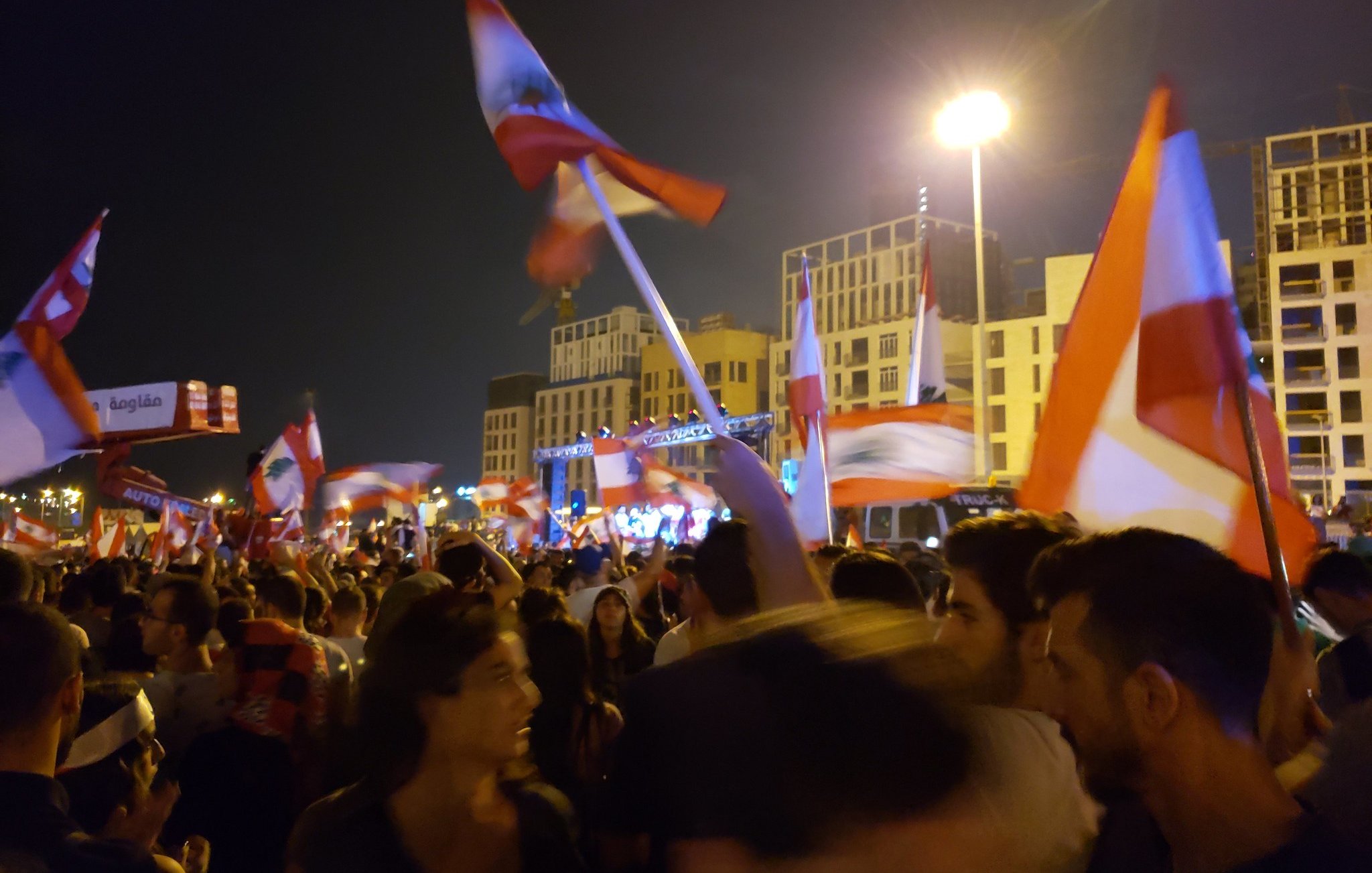 L'image montre une foule rassemblée à la nuit tombée, tenant des drapeaux libanais. Les gens semblent être en plein mouvement, probablement en train de célébrer ou de manifester. On aperçoit une scène à l'arrière-plan, éclairée par des lumières, ce qui suggère un événement festif ou politique. L'atmosphère est dynamique, remplie d'énergie, avec des expressions de joie et d'engagement parmi les participants.