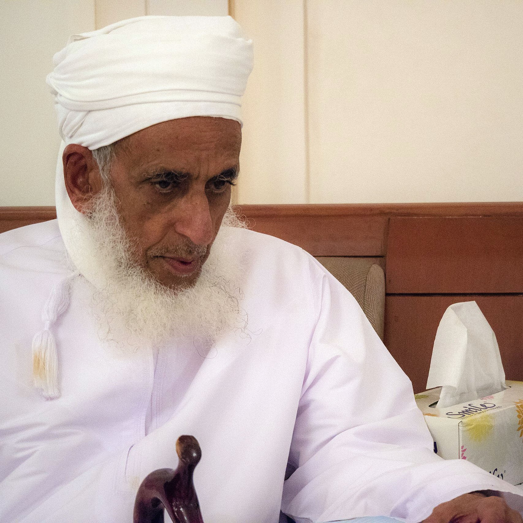 L'image montre un homme âgé portant un turban blanc et une robe traditionnelle. Il a une barbe grise et semble pensif, assis sur un canapé. À côté de lui, on peut voir des mouchoirs et un sac en papier, ce qui suggère qu'il se trouve dans un environnement intérieur calme. L'éclairage est doux, créant une atmosphère sereine.