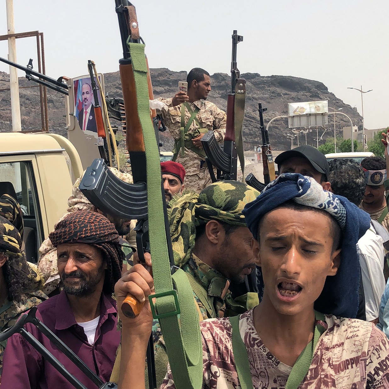 The image depicts a group of armed individuals standing together in an outdoor setting. They are dressed in military-style clothing and some wear traditional headscarves. Several of them are holding rifles and appear to be engaged in a rally or demonstration. The background features a vehicle and a large rock formation, with a hint of a cityscape. A flag is visible, adding to the atmosphere of the gathering, which seems to pertain to a political or military context. The expressions and postures of the individuals reflect a sense of determination or unity.