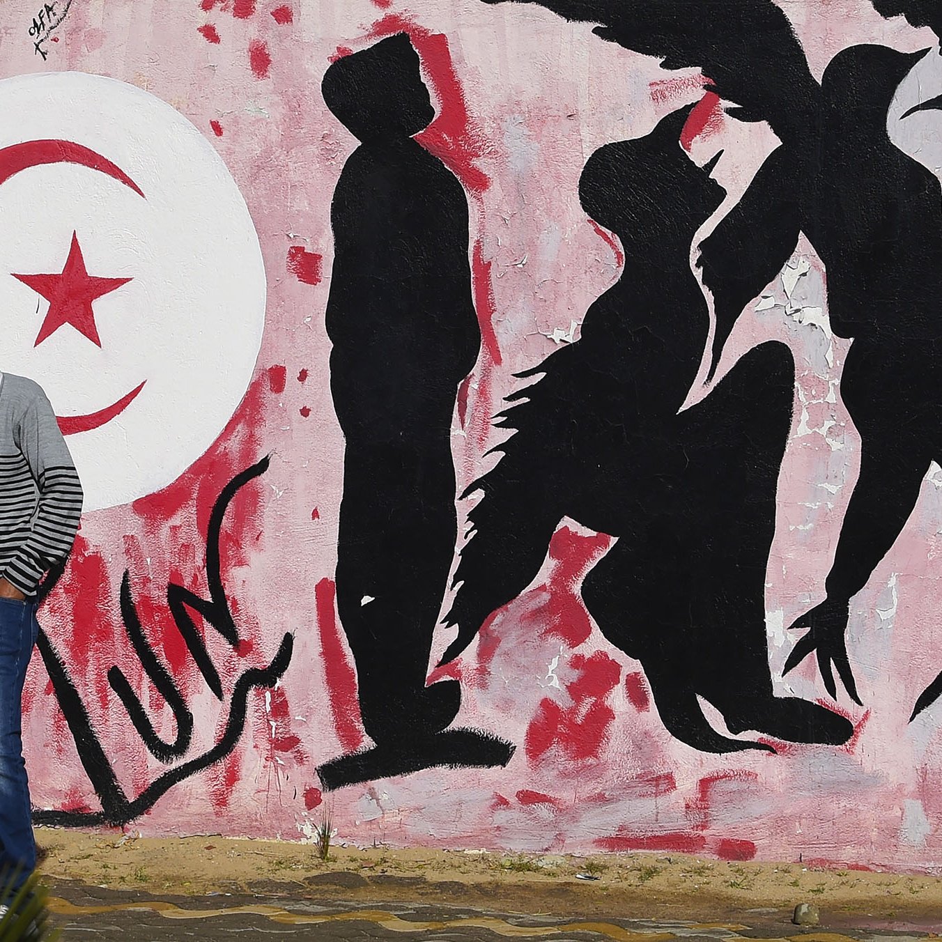 L'image montre un mur peint avec des graffitis. À gauche, on peut voir le drapeau tunisien en rouge et blanc, avec un croissant et une étoile. À côté, des silhouettes humaines sont représentées en noir sur un fond rose, certaines semblent interagir avec des éléments d'oiseaux, suggérant des thèmes de liberté ou de mouvement. Un homme en pull rayé marche le long du mur, ajoutant un aspect vivant à la scène.