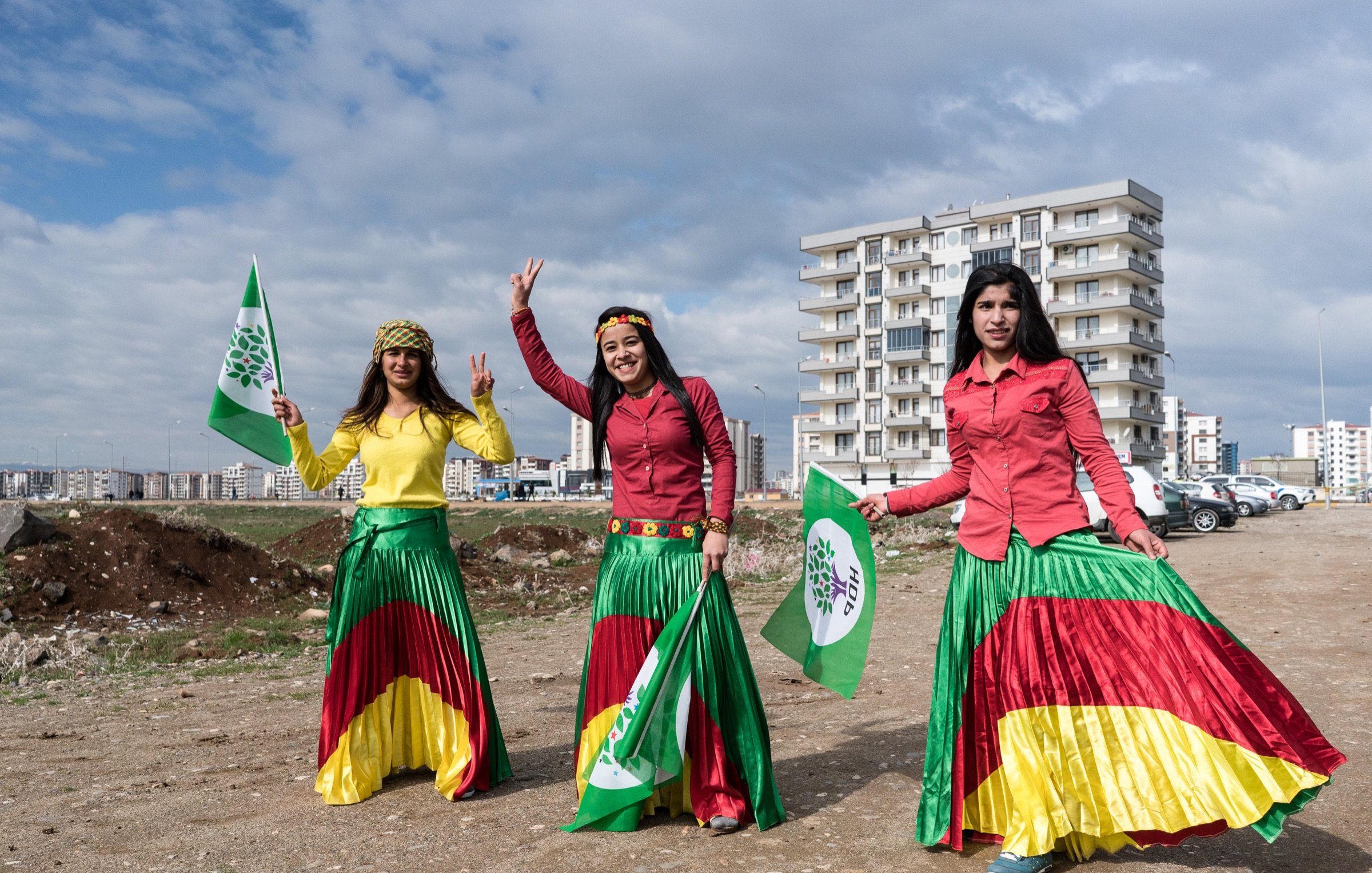 L'image montre trois femmes portant des jupes colorées, affichant des drapeaux et symboles. Elles sont entourées d'un paysage urbain, avec des bâtiments en arrière-plan et un terrain dégagé. Les femmes semblent célébrer un événement, affichant des sourires et des gestes de victoire. Les couleurs vives de leurs vêtements et des drapeaux évoquent un sentiment de fierté culturelle.