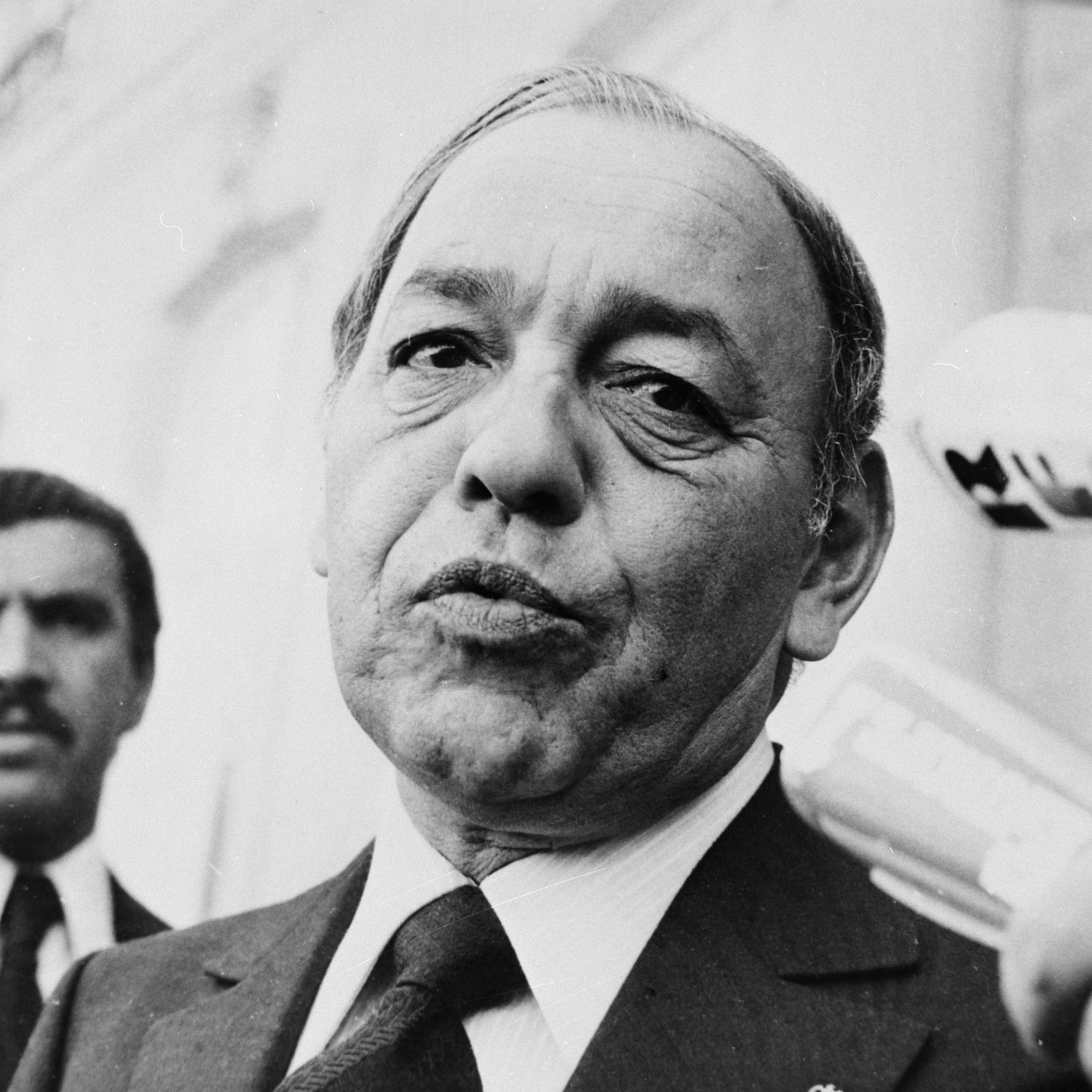 این تصویر یک مرد میانسال را نشان می‌دهد که ظاهری جدی و متمرکز دارد. او لباس رسمی به تن دارد و به دوربین نگاه می‌کند. در پس‌زمینه، شخص دیگری نیز دیده می‌شود که به نظر می‌رسد در حال نظارت یا همراهی با او است. فضای تصویر به طور کلی رسمی و مرتبط با یک موقعیت اجتماعی یا سیاسی به نظر می‌رسد.