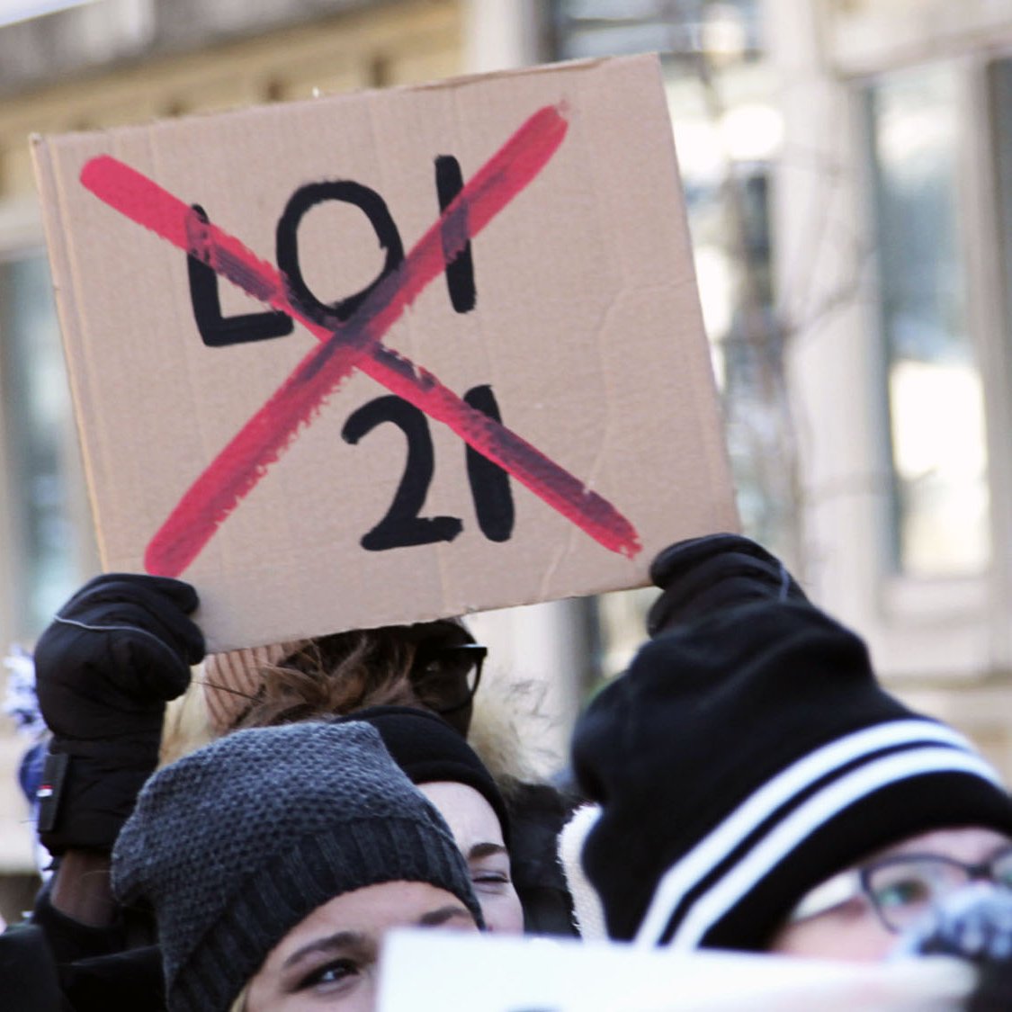 The image depicts a group of people participating in a protest. One prominent sign held up by a protester is made of cardboard and has "LOI 21" (with a red "X" crossing it out) written in bold letters. The crowd appears to be wearing winter clothing, such as hats and gloves, indicating that the protest is taking place in a cold environment. The atmosphere seems to be one of solidarity and activism, as the participants are visibly engaged in the demonstration.