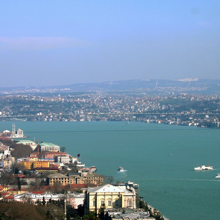 L'image montre un panorama d'une ville au bord d'une grande étendue d'eau. On aperçoit un pont moderne qui traverse la rivière, reliant les deux rives. À gauche, des bâtiments s'échelonnent sur la colline, tandis qu'à droite, on peut voir des constructions historiques. Le ciel est clair, et l'ensemble du paysage évoque une atmosphère tranquille et pittoresque. On distingue également des bateaux naviguant sur la rivière.