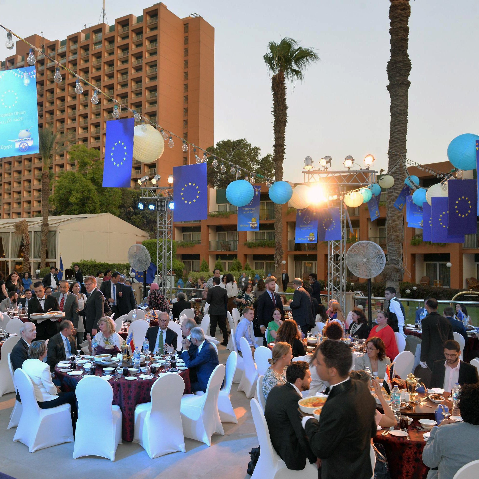 الصورة تظهر احتفالًا أو مناسبة اجتماعية في الهواء الطلق. هناك طاولات مزينة بأقمشة ملونة، وحولها مجموعة من الناس يرتدون ملابس أنيقة. في الخلفية، يبرز مبنى مرتفع، ويحيط بالمنطقة أشجار النخيل. الأضواء الداخلية والخارجية تعزز الأجواء الاحتفالية، وتوجد زينة على شكل كرات ملونة، بالإضافة إلى لافتات تحمل شعار الاتحاد الأوروبي. الأجواء تبدو مفعمة بالحياة والهدوء في الوقت نفسه.