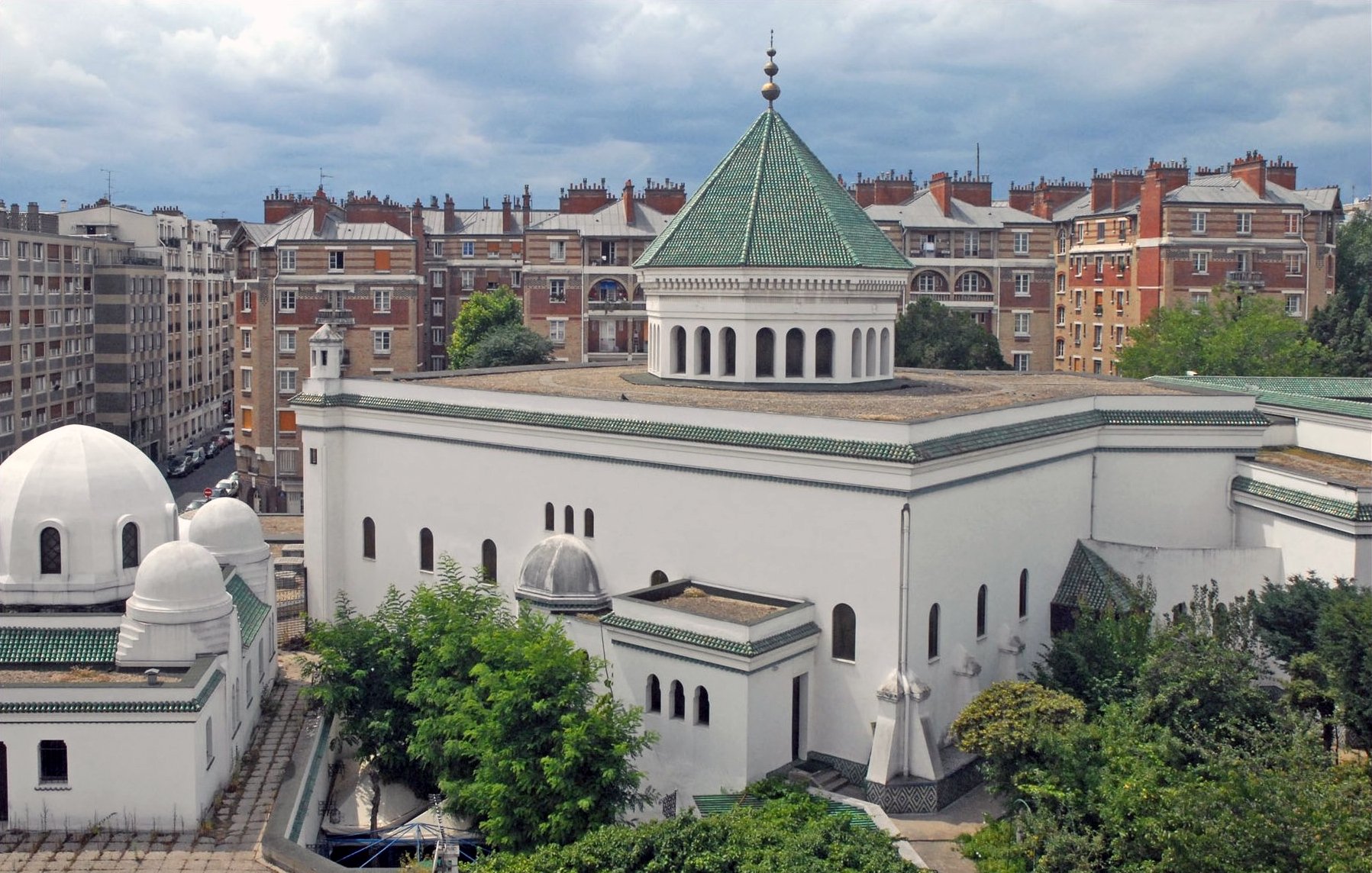 L'image montre un bâtiment architectural distinctif avec une façade blanche et un toit en tuiles vertes. Le design présente des éléments inspirés de l'architecture islamique, comme des dômes et des arches. En arrière-plan, on aperçoit des immeubles de style haussmannien, typiques de nombreuses villes françaises, avec des balcons et des toits en attique. Le ciel est nuageux, ce qui ajoute une atmosphère particulière à la scène. Des arbres et de la verdure entourent le bâtiment, contribuant à l'esthétique générale.