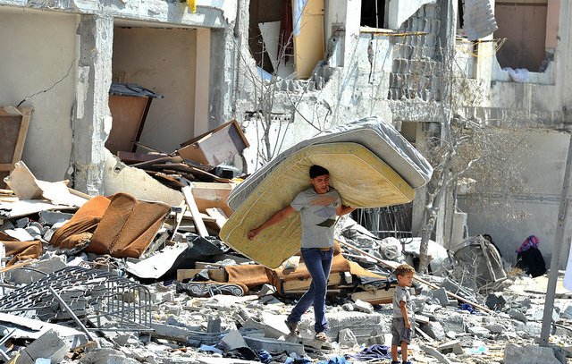 L'image montre un jeune homme portant un matelas sur son dos, marchant à travers des décombres. À côté de lui, un petit garçon l'observe. Le décor est celui d'un bâtiment en ruines, avec des meubles éparpillés et des murs endommagés, ce qui suggère une scène de destruction et de détresse. L'atmosphère est empreinte de tristesse et de désolation.