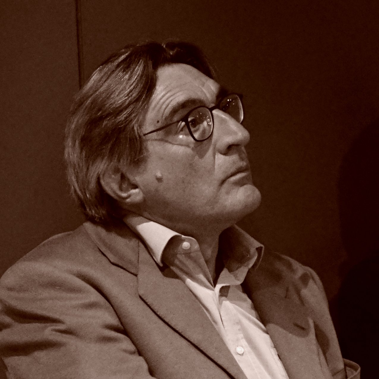 L'image montre un homme aux cheveux grisonnants, portant des lunettes et un costume clair. Il semble pensif, regardant quelque chose hors champ. L'éclairage et le ton de l'image lui donnent un aspect rétro, suggérant une ambiance contemplative.