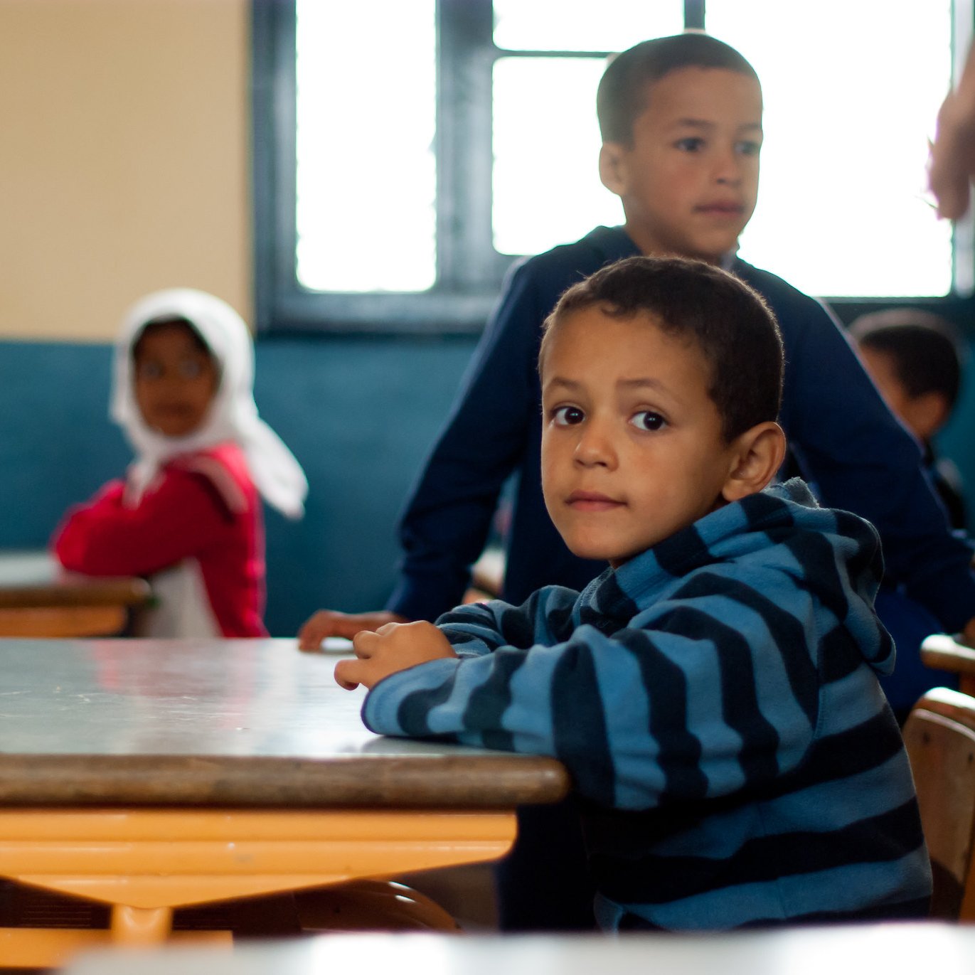 L'image montre une salle de classe où plusieurs enfants sont présents. Au premier plan, un jeune garçon avec un pull à rayures bleues et noires est assis à un bureau, regardant vers l'objectif avec une expression curieuse. À l'arrière-plan, on aperçoit d'autres enfants, dont un garçon debout et une fille avec un foulard. Les murs de la classe sont peints en bleu, et la lumière entre par des fenêtres. L'atmosphère semble calme et concentrée.