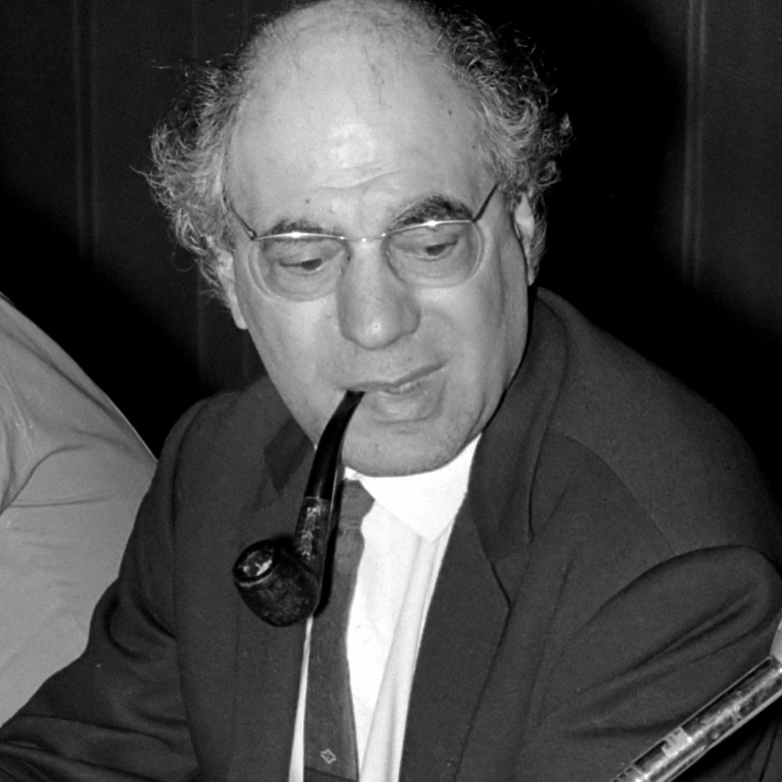 L'image montre un homme assis avec une pipe dans la bouche. Il porte une veste, une chemise blanche et une cravate. Ses cheveux sont grisonnants et légèrement bouclés. Il a des lunettes et semble concentré sur une conversation ou une discussion. En arrière-plan, on peut apercevoir d'autres personnes, mais elles ne sont pas clairement visibles. L'éclairage est modéré, ce qui donne une ambiance sérieuse à la scène.