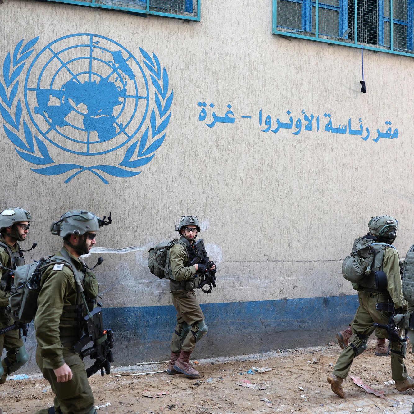 تظهر الصورة مجموعة من الجنود يرتدون زيًا عسكريًا ويقومون بالتحرك في منطقة قريبة من مبنى يحمل شعار الأمم المتحدة باللغة العربية. الجدران تحمل كتابة تشير إلى مقر الأونروا في غزة، ويبدو أن المكان يعاني من بعض الفوضى أو الأضرار. الجنود يظهرون وكأنهم في حالة تأهب، مما يعكس أجواء من التوتر أو النشاط العسكري في المنطقة.