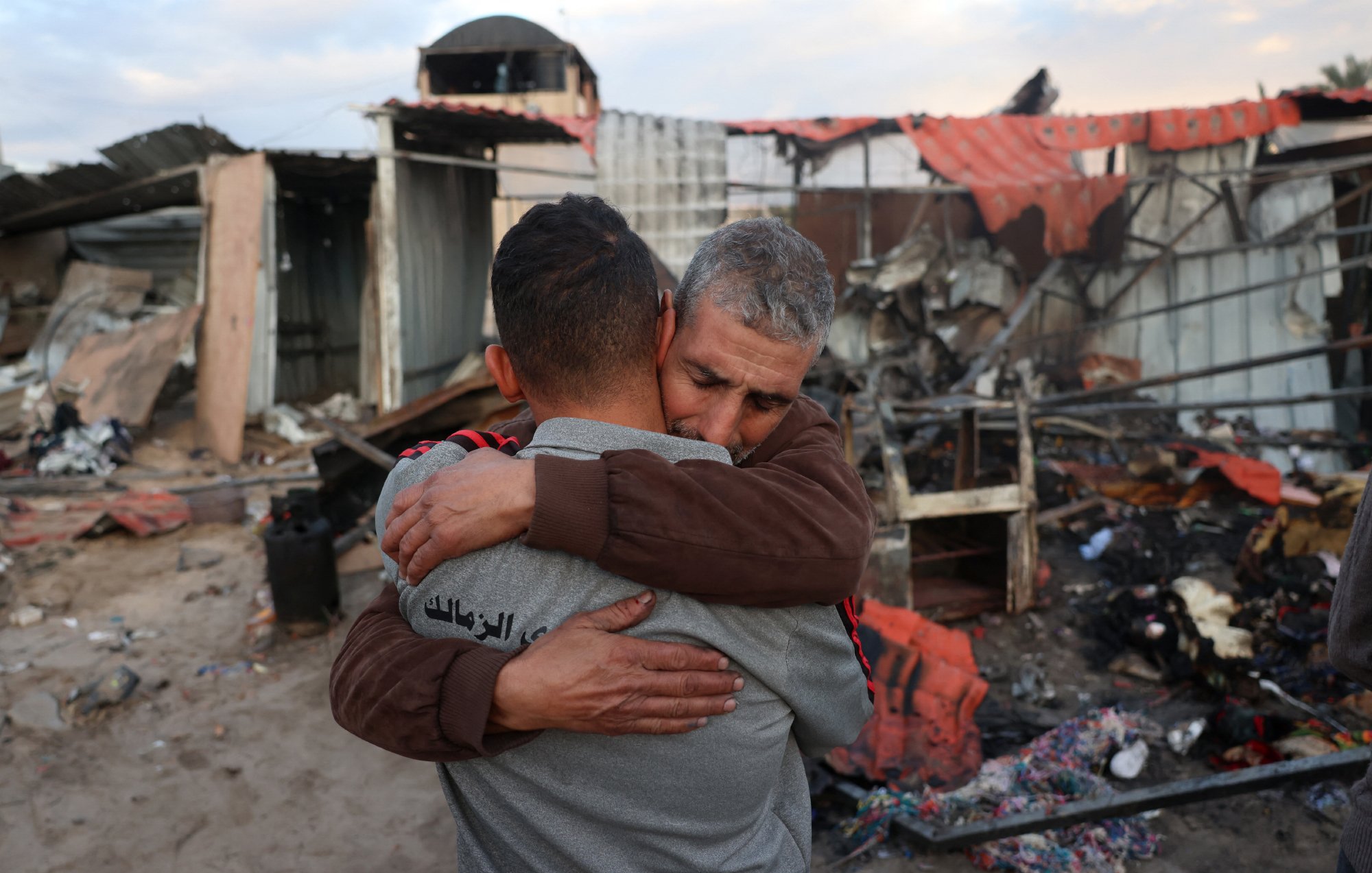 در این تصویر، دو مرد به یکدیگر در آغوش می‌افتند. پس‌زمینه تصویر نشان‌دهنده یک محل ویران شده است که به نظر می‌رسد دچار آتش‌سوزی یا تخریب شده است. حس ناراحتی و همدردی در چهره‌های آنها مشهود است و فضای کلی تصویر احساساتی و غم‌انگیز به نظر می‌رسد.