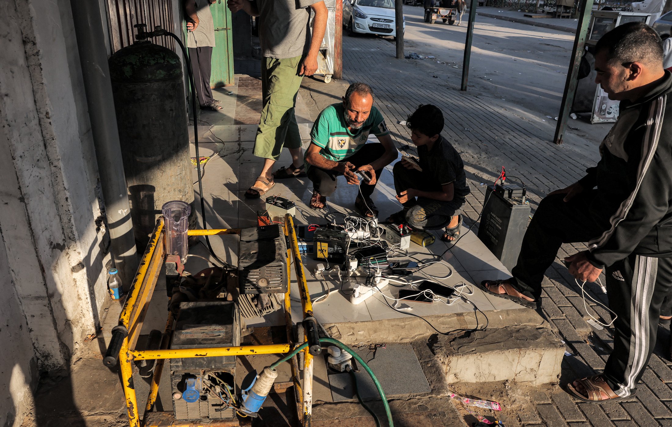 Nell'immagine si possono vedere alcune persone raccolte in un'area urbana. Tre uomini sono intenti a lavorare attorno a un dispositivo, probabilmente un generatore elettrico, che è posizionato su una base di cemento. Accanto al generatore ci sono vari cavi e dispositivi elettronici, suggerendo che stanno cercando di ricaricare batterie o alimentare apparecchi. Uno degli uomini è accovacciato mentre gli altri due stanno osservando o interagendo con i cavi. Sullo sfondo, si intravedono auto e strutture urbane tipiche. L'atmosfera sembra essere di lavoro e collaborazione nel contesto di un'area pubblica.
