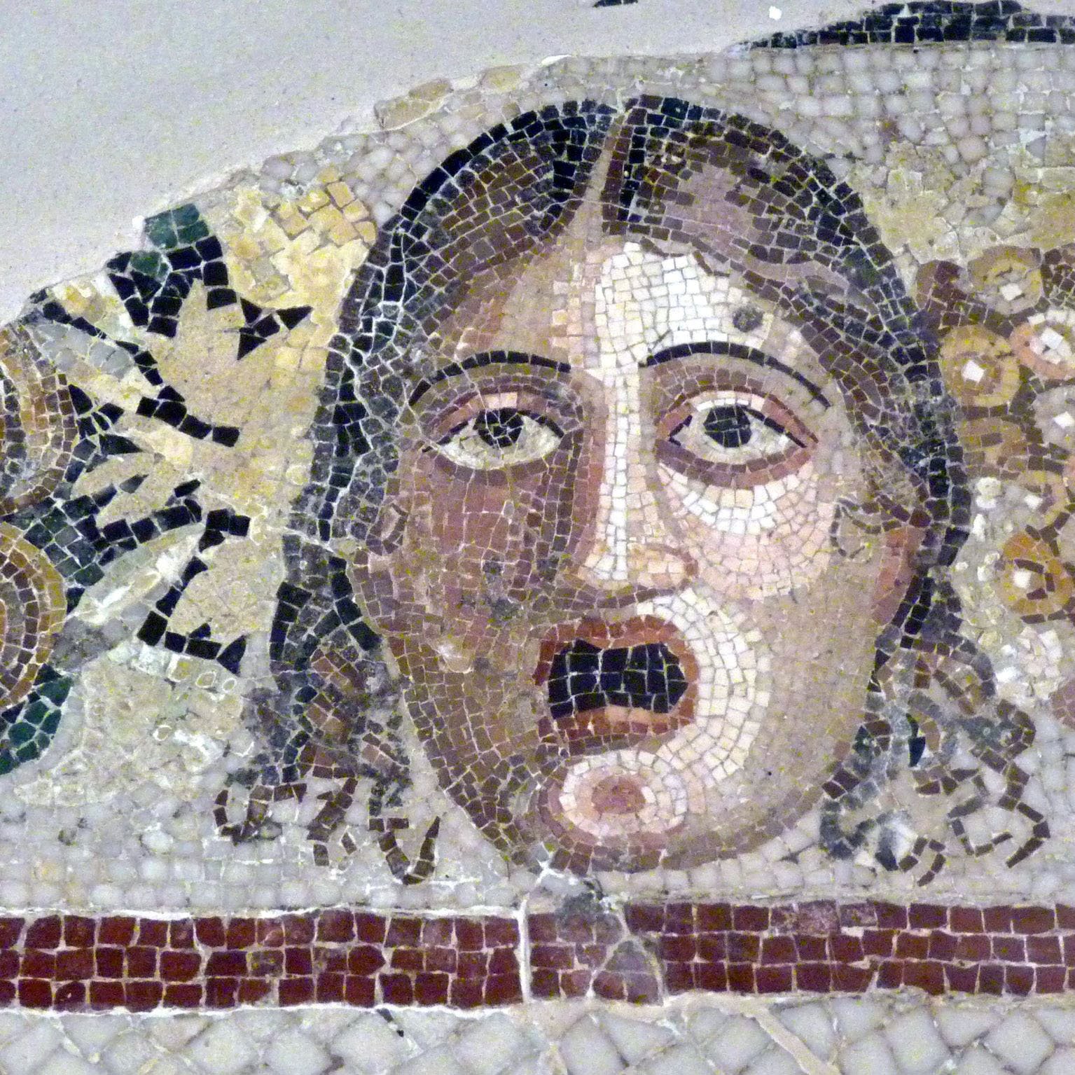 The image depicts a detailed mosaic featuring a face with a startled or shocked expression. The individual's wide eyes and open mouth convey a sense of surprise or fear. Surrounding the face are decorative elements, including clusters of grapes and possibly other fruits or foliage, which suggest a theme of abundance or festivity. The mosaic is composed of various colored tiles, showcasing intricate craftsmanship typical of ancient art.