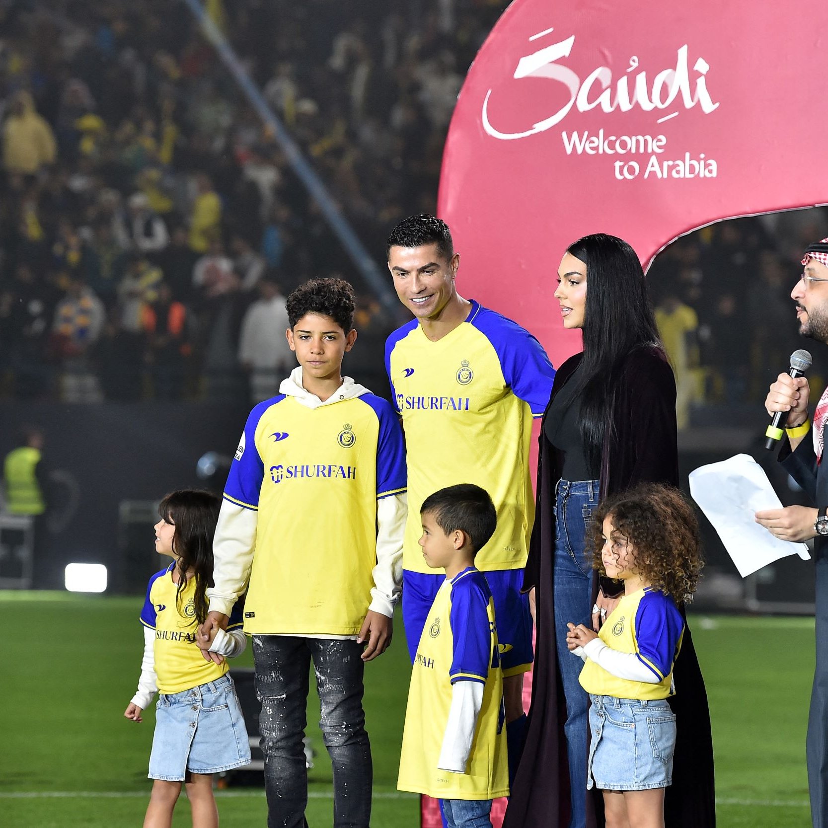 L'image montre une scène sur un terrain de football, où plusieurs personnes sont réunies autour d'un joueur, probablement un footballeur célèbre, en tenue de jeu. Il y a des enfants, probablement des membres de la famille, qui se tiennent à ses côtés. Une femme, habillée d'une manière élégante, semble s'adresser à l'audience, tandis qu'un homme en tenue traditionnelle parle également en tenant un microphone. En arrière-plan, des spectateurs sont visibles, et une grande arche avec les mots "Welcome to Arabia" est installée, indiquant un événement spécial.