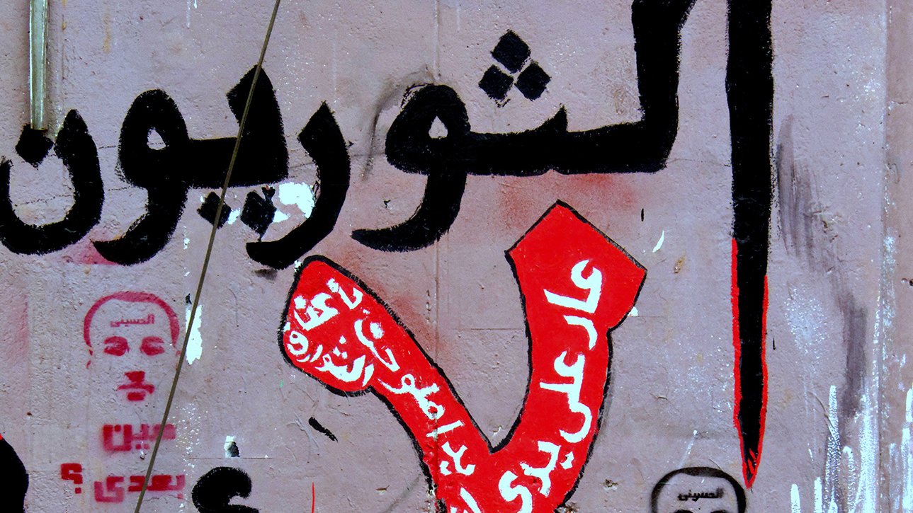 L'image montre un mur recouvert de graffitis, avec des inscriptions en arabe. On peut y voir le mot "Non" stylisé en rouge, entouré de phrases opposées à certaines idées ou actions. Des visages ou portraits, probablement des figures publiques, sont également esquissés. L'ensemble de la composition véhicule un message fort et engagé, typique de la communication visuelle dans des contextes de protestation sociale ou politique.