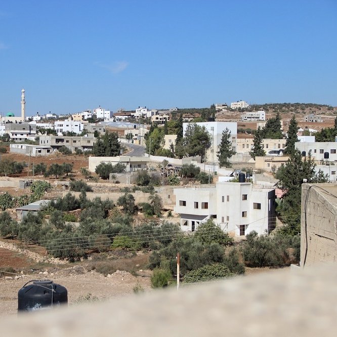 L'image montre un paysage urbain de type rural, avec des maisons de couleur claire disséminées sur une colline. On peut apercevoir des arbres et des champs, ainsi qu'un minaret se dressant dans le fond, ce qui pourrait indiquer la présence d'une mosquée. Le ciel est dégagé et d'un bleu vif, ce qui donne à la scène une atmosphère ensoleillée. Les infrastructures semblent simples et typiques des zones périphériques.