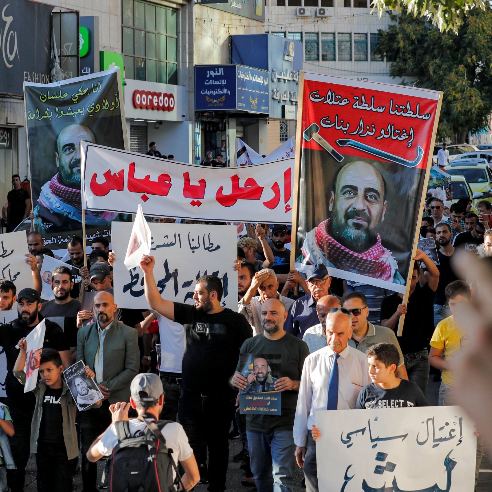 L'image montre une foule de manifestants dans une rue. Les participants portent des pancartes et des banderoles avec des écrits en arabe. L'ambiance semble être celle d'une manifestation ou d'un rassemblement public. Plusieurs personnes brandissent des affiches, et on peut voir des voitures et des bâtiments en arrière-plan. Les manifestants semblent engagés et déterminés dans leur action.