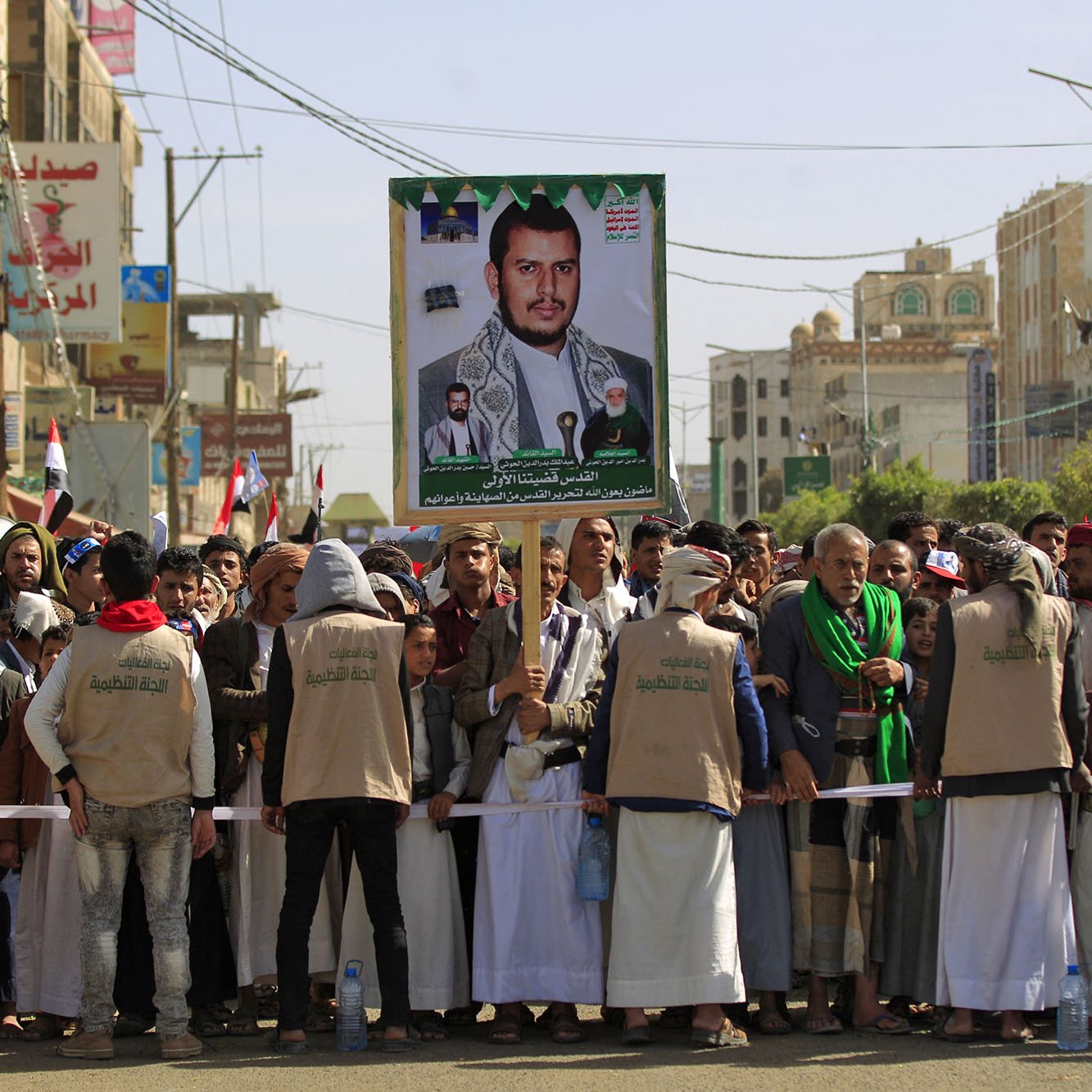 تظهر في الصورة مجموعة من الأشخاص مجتمعين في مظاهرة أو تجمّع شعبي. في الخلفية، يُرفع لافتة كبيرة تحمل صورة لأحد الشخصيات، ويبدو أن المتظاهرين يرتدون ملابس متشابهة. تعكس الأعلام الموجودة في الصورة انتماءات وطنية، كما يظهر أن هناك تفاعلًا وحماسًا بين المشاركين. الجو يعكس نوعًا من الاحتشاد والتضامن حول قضية معينة.