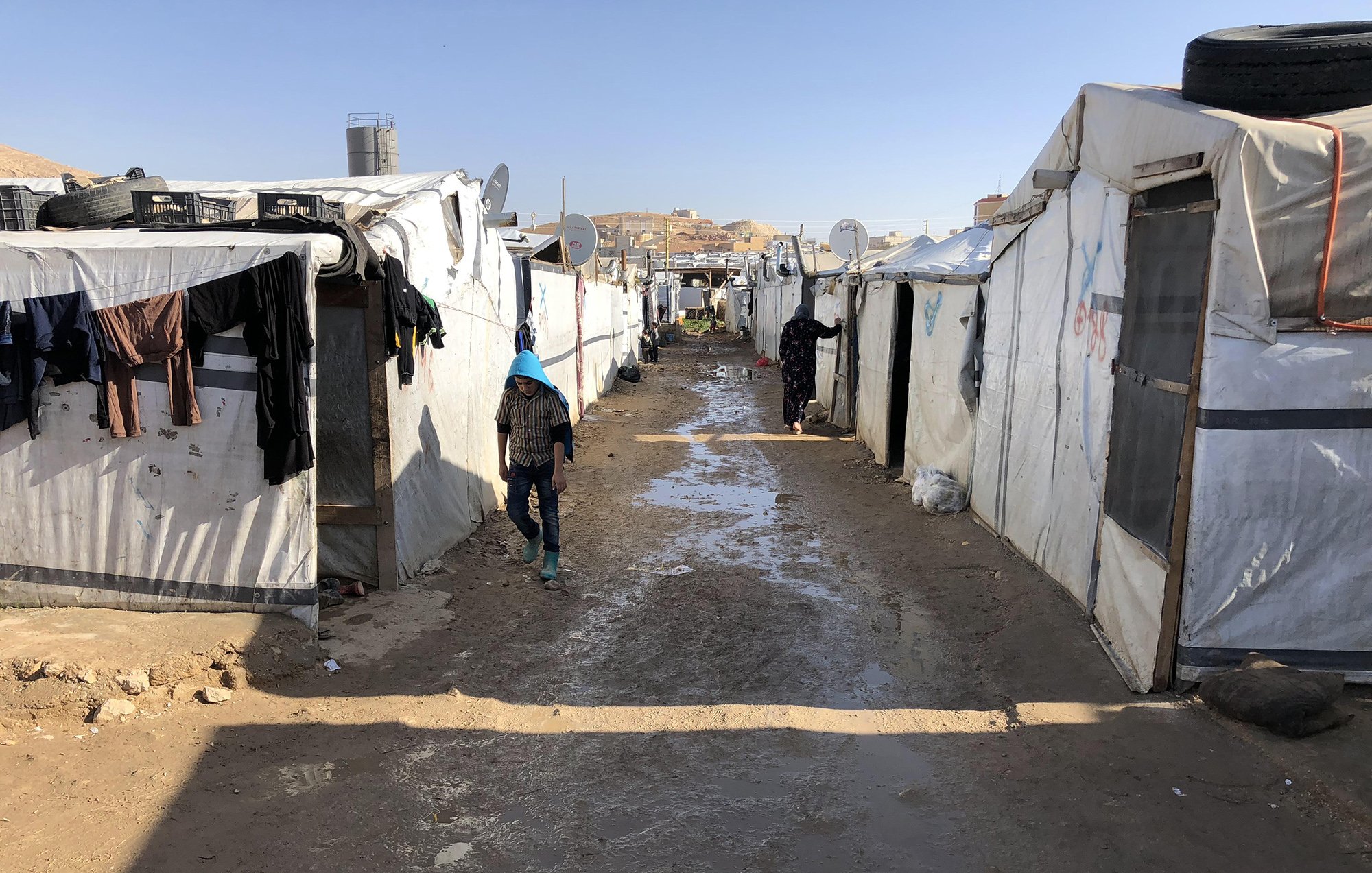 این تصویر نمایی از یک کمپ پناهندگان را نشان می‌دهد. در اینجا، چادرهای سفید و مشکی به صورت ردیفی در دو طرف یک مسیر خاکی قرار دارند. زمین به نظر می‌رسد کمی مرطوب و گل‌آلود است. چندین قطعه لباس بر روی چادرها آویزان شده‌اند و شخصی در حال حرکت در مسیر دیده می‌شود. در پس‌زمینه، ساختمان‌هایی در بالای تپه‌ها قابل مشاهده هستند. این منظره فضایی ساده و دلگیر را تداعی می‌کند.