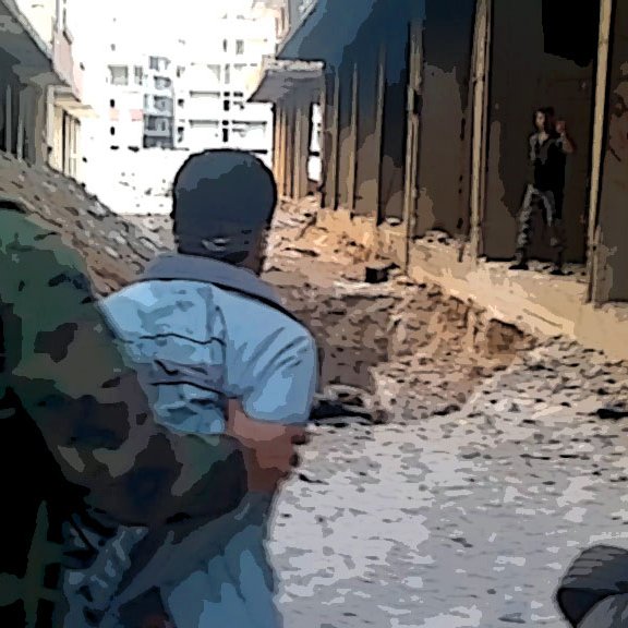 The image depicts a war-torn urban environment. A soldier in military attire is seen guiding or restraining a person wearing a light-colored shirt. The setting suggests destruction, with debris scattered on the ground and the remnants of buildings in disrepair. In the background, another individual is partially visible, possibly further down the street. The overall atmosphere conveys a sense of tension and conflict.