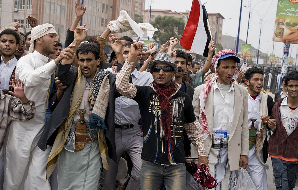 L'image montre un groupe de personnes en train de manifester dans une rue. Elles semblent exprimer des revendications, levant les bras et brandissant des drapeaux, notamment un drapeau rouge. Les manifestants portent des vêtements traditionnels yéménites, avec divers styles de vêtements. L'ambiance paraît énergique et engagée, comme une expression de protestation ou de soutien à une cause.