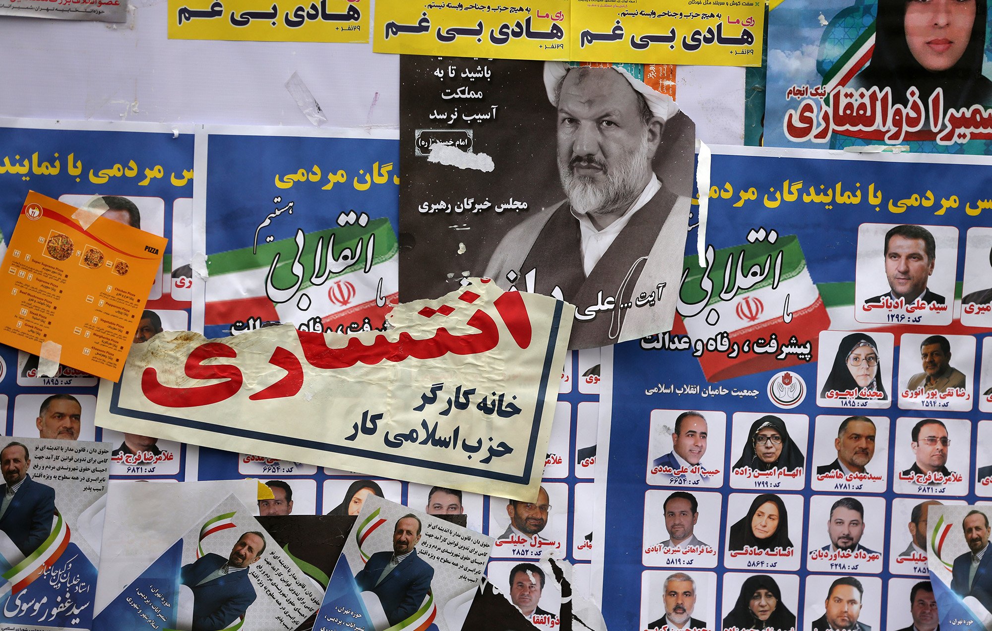 L'image montre un mur recouvert de diverses affiches, probablement liées à des élections. On peut y voir des portraits de candidats, des slogans politiques en farsi, et des éléments graphiques colorés. Certaines affiches semblent promouvoir des partis politiques ou des mouvements, tandis que d'autres affichent des messages mobilisateurs pour inciter à voter. L'ensemble crée une atmosphère vibrante et engageante, typique des campagnes électorales.