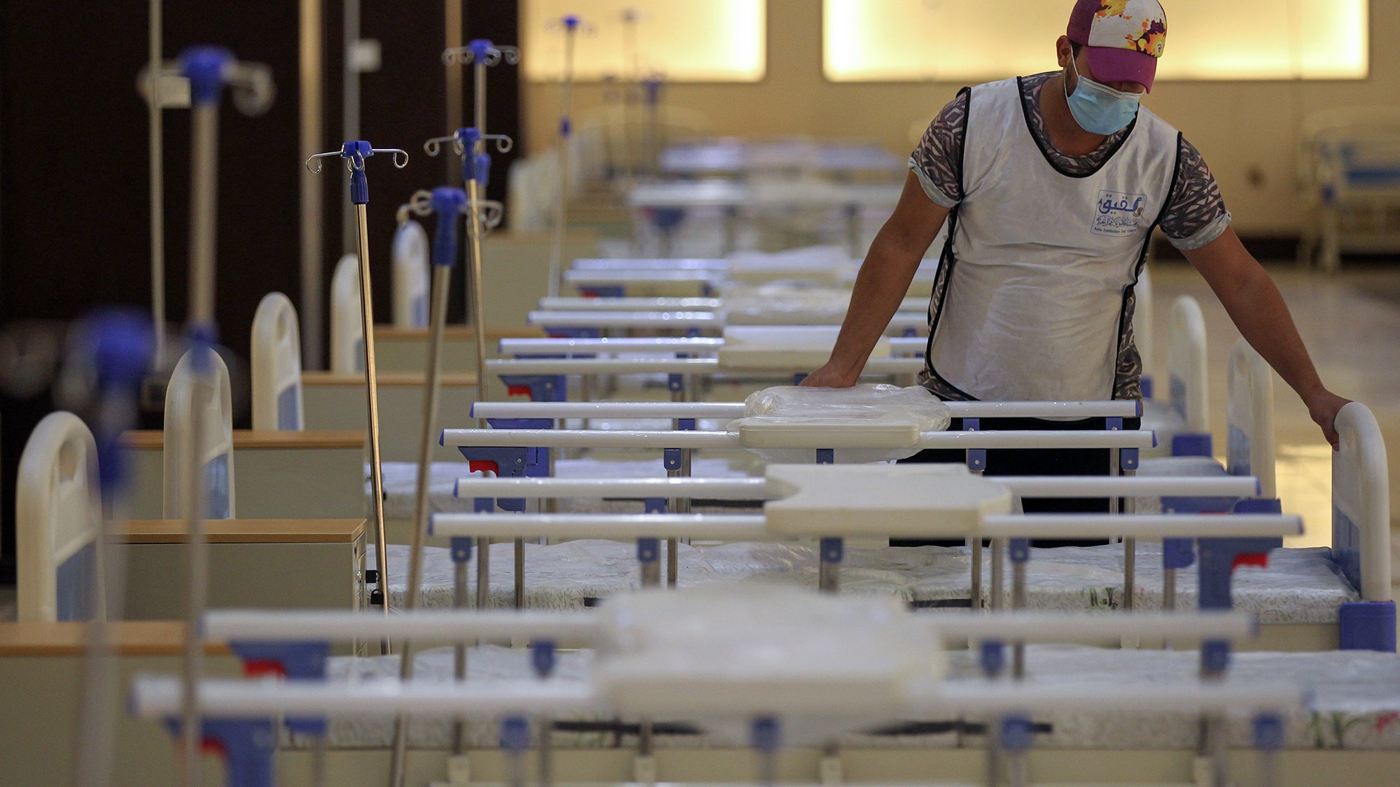 La imagen muestra a un hombre que está de pie junto a una serie de camas dispuestas en una sala. El hombre lleva una mascarilla y un chaleco, mientras que las camas están organizadas en filas, con sábanas y equipos médicos visibles. La iluminación es suave, lo que sugiere un ambiente de cuidado hospitalario. La escena da la impresión de un espacio preparado para atender pacientes.