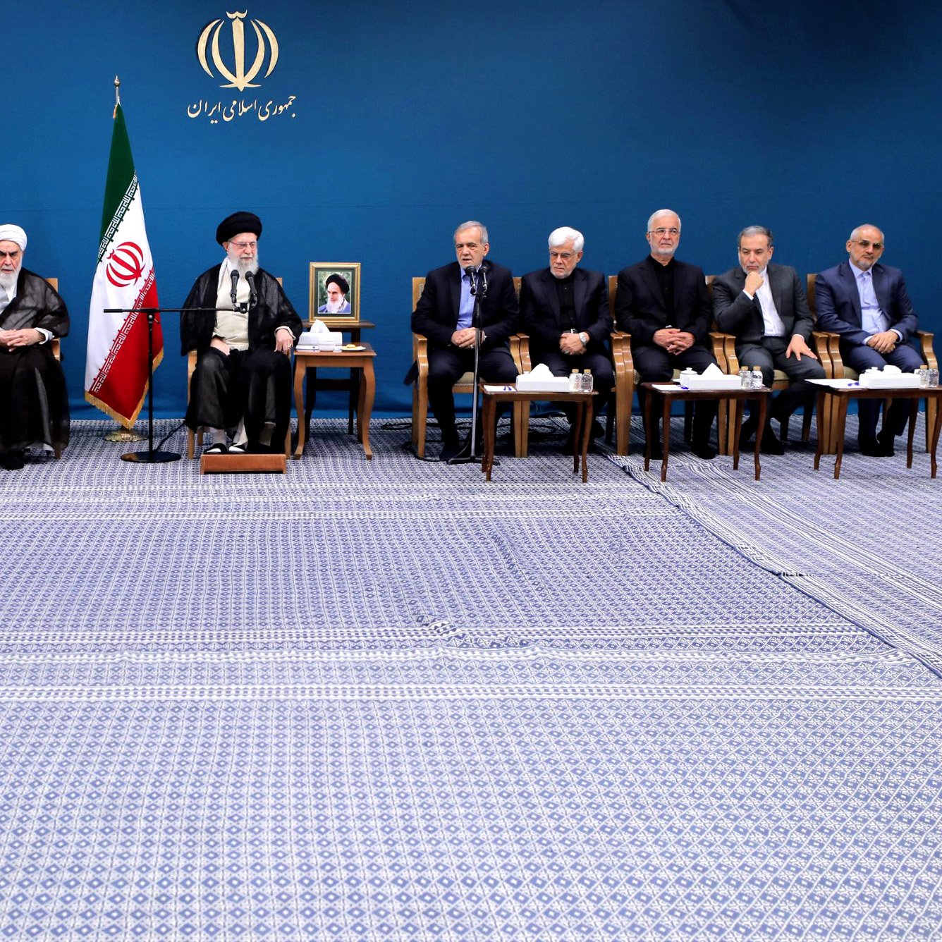 این تصویر نشان‌دهنده یک نشست یا جلسه در ایران است. در پس‌زمینه، پرچم ایران و تصویری از یک مقام ارشد دیده می‌شود. چندین فرد به صورت گروهی نشسته‌اند، برخی با لباس رسمی و دیگران با لباس‌های سنتی. به نظر می‌رسد که این افراد شامل مقامات دولتی و دیگر شخصیت‌های مهم کشور باشند. فضای اتاق به‌طور سنتی و رسمی چیده شده و فرش بزرگی زیر پای آن‌ها پهن شده است.