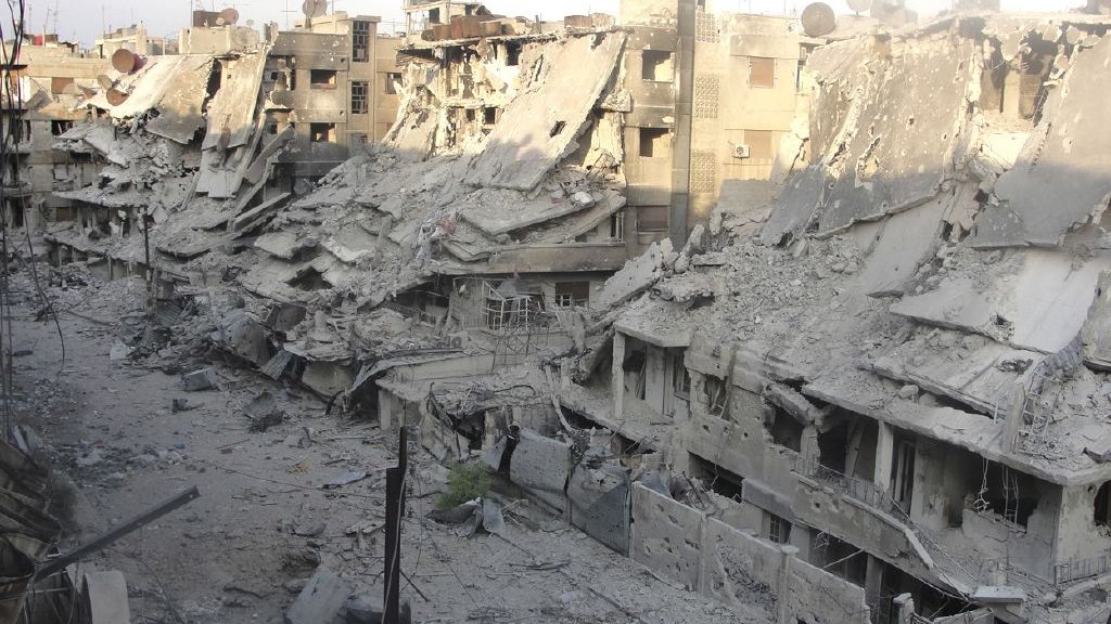 L'image montre un paysage urbain fortement endommagé, avec des bâtiments effondrés et en ruines. Les façades des immeubles sont détruites, laissant entrevoir des débris et des structures instables. Le sol est recouvert de débris et de poussière, témoignant de la violence des destructions. La lumière du jour éclaire la scène, accentuant le contraste entre les ruines et le ciel. Cette image évoque un environnement de guerre et de crise humanitaire.