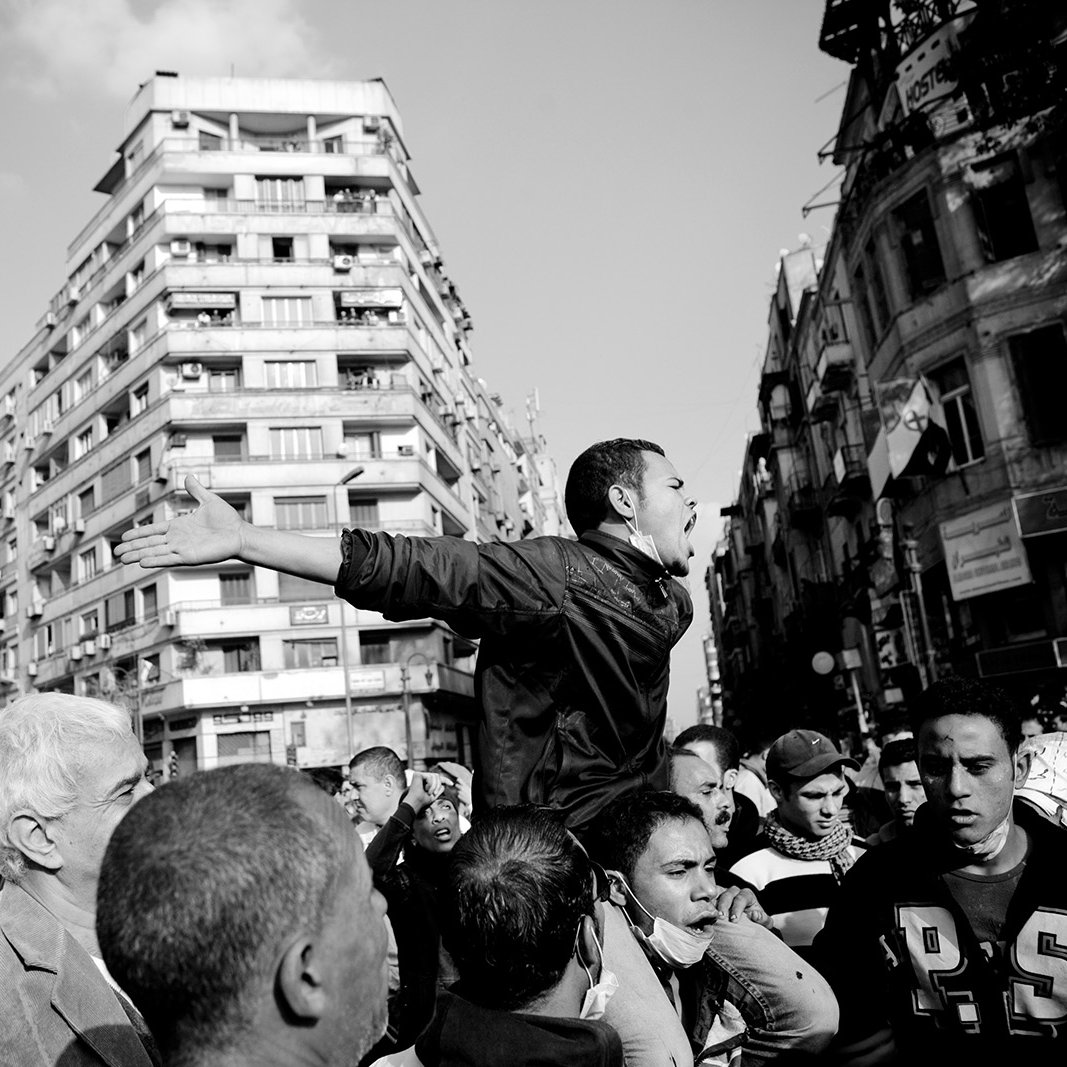 تظهر الصورة مشهدًا لحشود من الناس في منطقة حضرية. في المقدمة، يوجد رجل يرفع يديه ويبدو أنه يتحدث أو يصرخ بوجه الآخرين. يحيط به عدد من الرجال الذين يظهرون تعابير مختلفة، مما يدل على تفاعلهم مع ما يقوله. الخلفية تحتوي على مباني شاهقة تعكس طابع المدينة. الجو العام يوحي بالتوتر أو الحماسة، مما يدل على وجود حدث مهم أو مظاهرة.