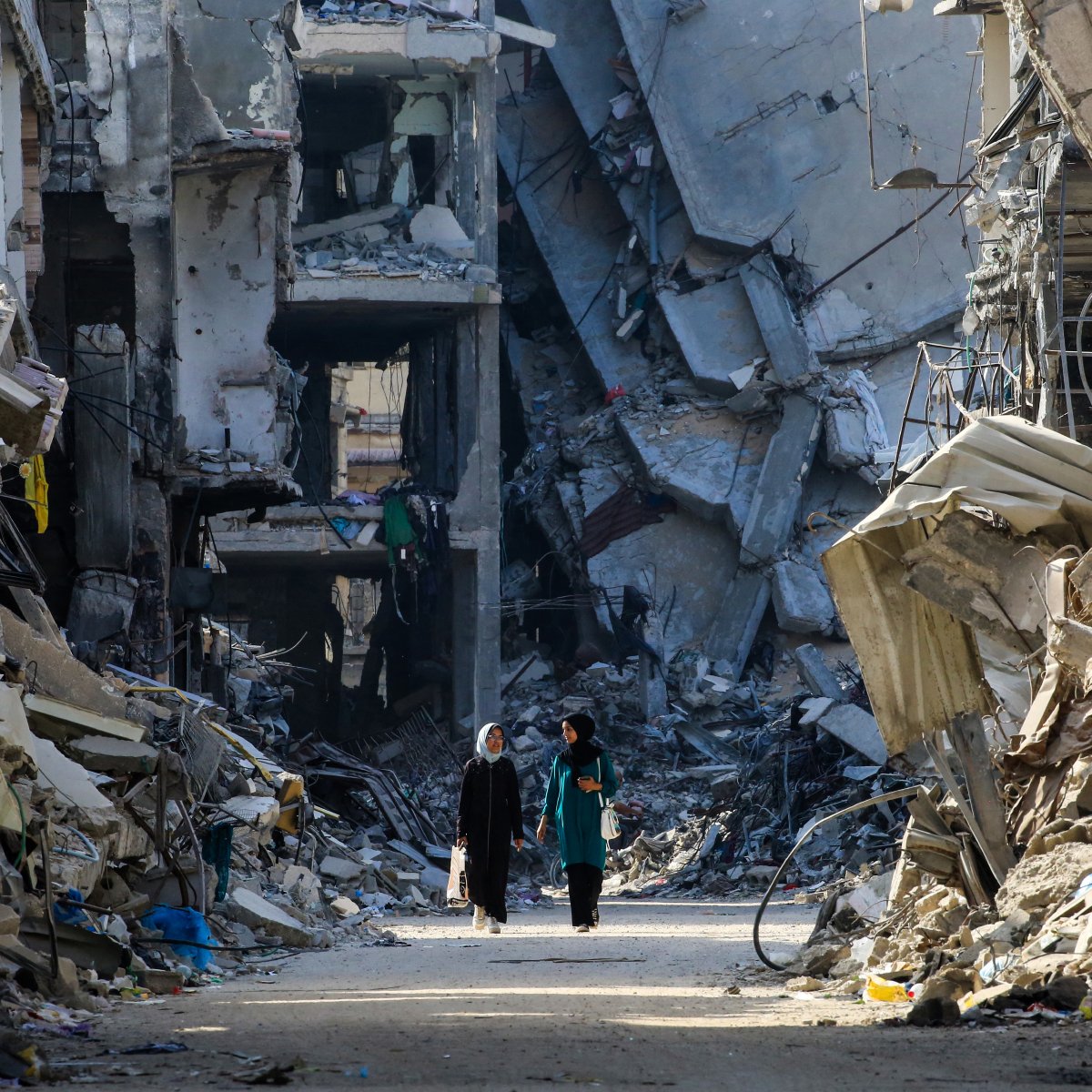 L'image montre un paysage de destruction urbaine, avec des bâtiments effondrés sur les côtés d'une rue. On peut apercevoir deux personnes marchant au milieu des décombres. L'atmosphère est sombre et désolée, avec des ruines qui témoignent d'une catastrophe récente. Les déchets et les restes de matériaux de construction jonchent le sol, créant un sentiment de désolation.