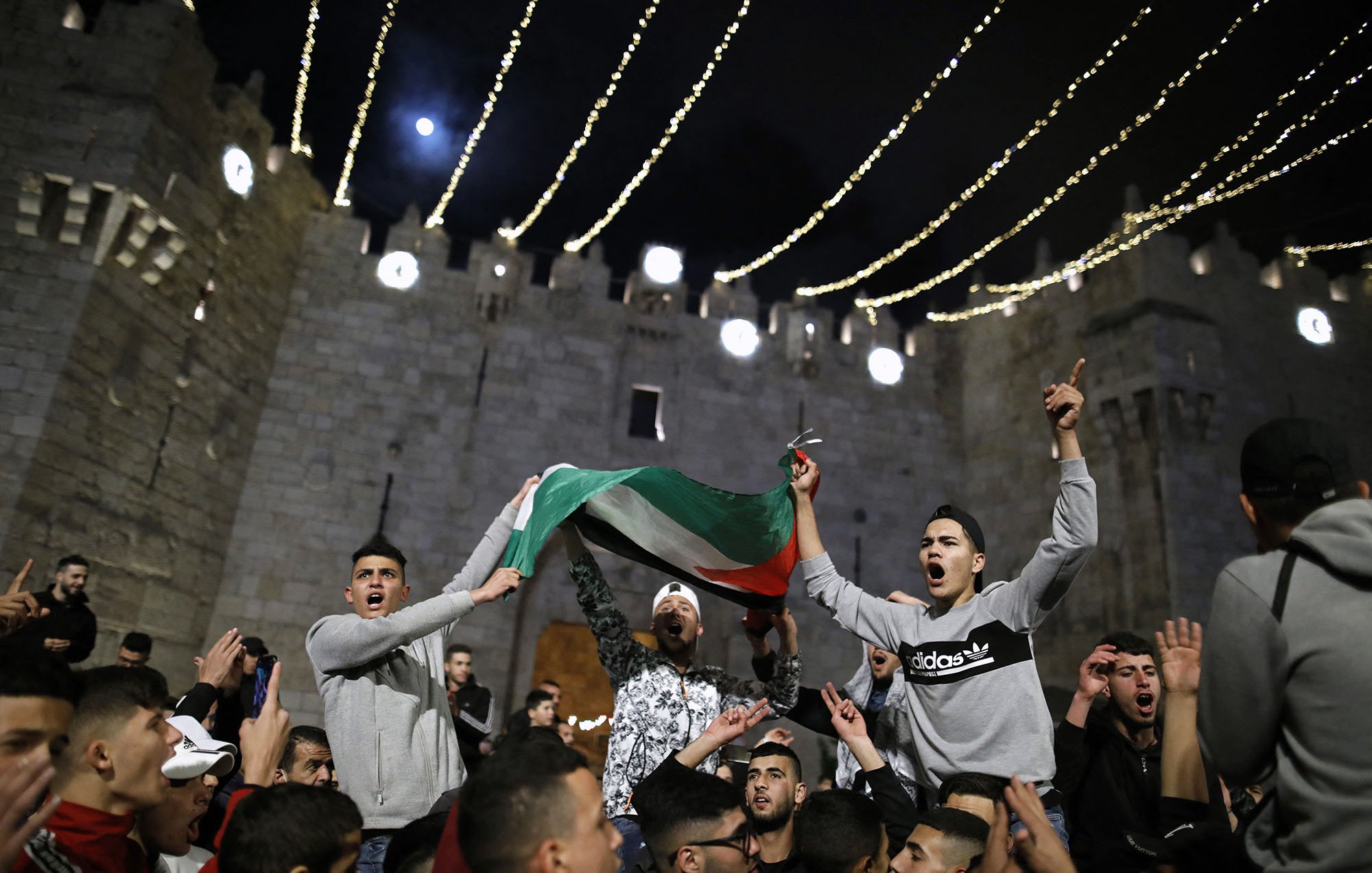 L'image montre un rassemblement nocturne devant un bâtiment ancien, probablement une fortification. Des personnes sont réunies en célébrant, certaines agitant un drapeau palestinien. L'atmosphère semble festive et dynamique, avec des lumières scintillantes qui illuminent la scène. Les expressions des participants retranscrivent une ambiance d'excitation et d'engagement.