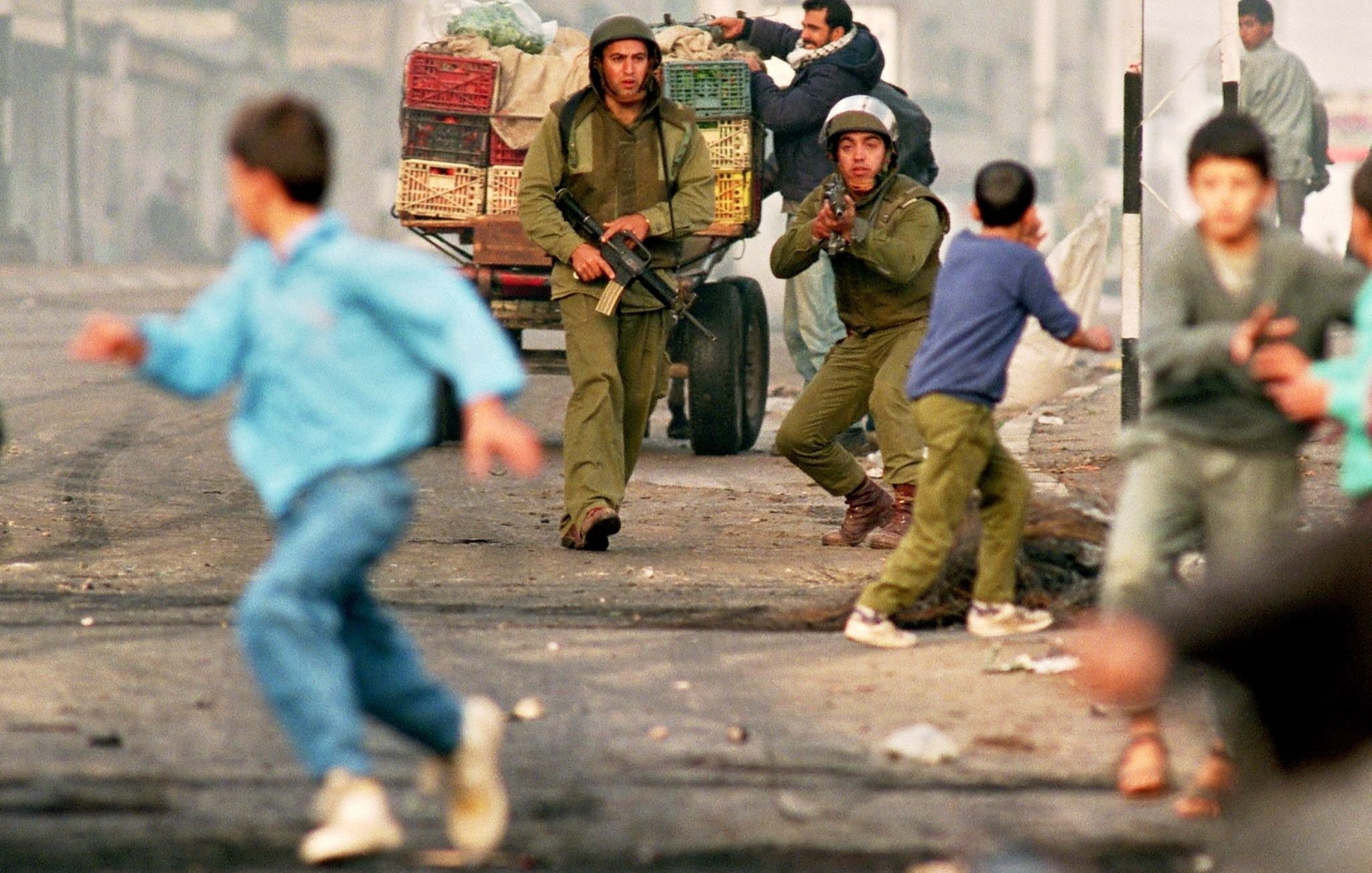 L'image montre une scène de tension dans un environnement urbain, où deux soldats sont présents, armés et en position d'action. Un homme, probablement un civil, est également visible, tandis que des enfants et d'autres personnes s'éloignent rapidement de la situation, suggérant un climat de peur ou de chaos. Des véhicules et des baraques sont en arrière-plan, accentuant l'atmosphère de conflit. L'ensemble évoque un contexte de violences ou d'affrontements dans une ville.