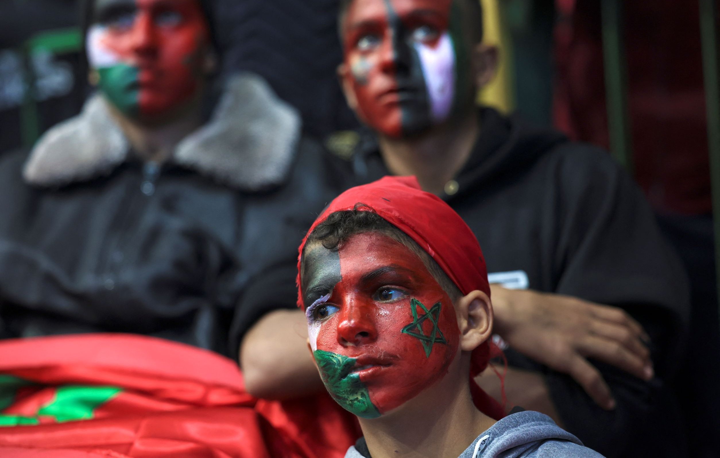 L'image montre un groupe de jeunes, dont un enfant au premier plan, qui ont le visage peint avec des motifs aux couleurs du drapeau marocain. L'enfant porte un bandana rouge et des dessins représentant le drapeau marocain et d'autres motifs sur son visage. En arrière-plan, deux autres personnes affichent également des visages peints, créant une atmosphère festive et engagée. L'ambiance semble liée à un événement de soutien ou à une célébration.