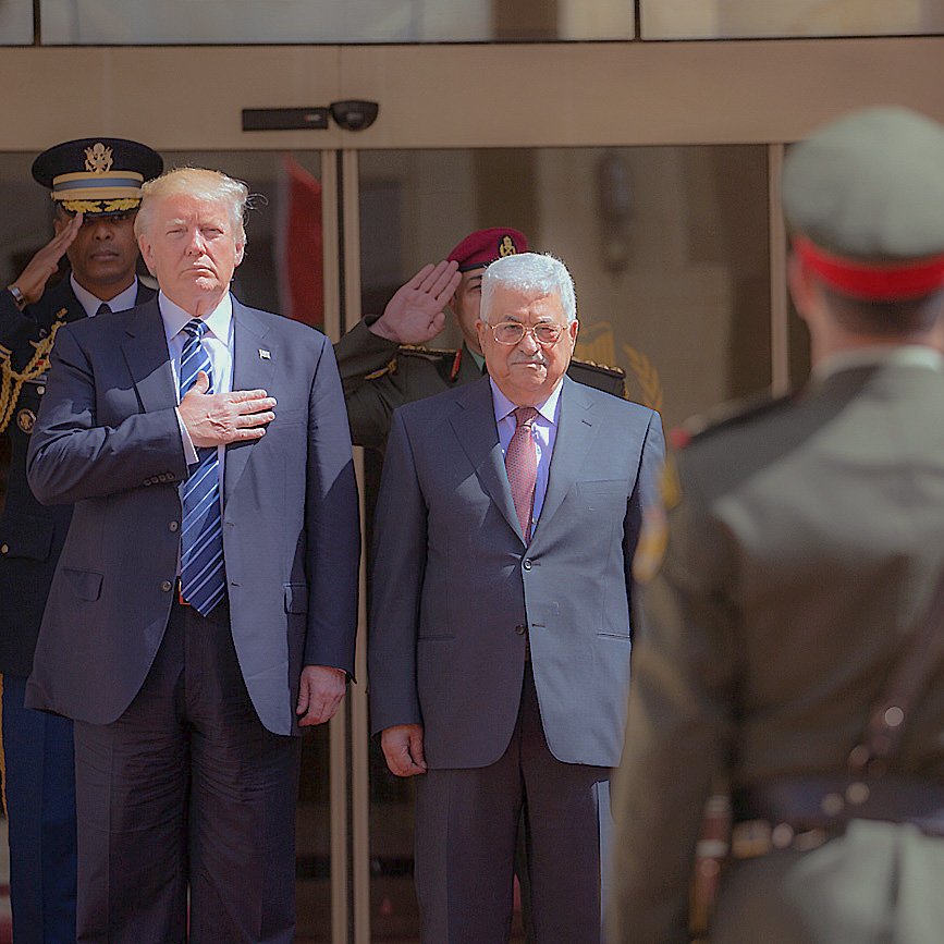 L'image montre deux hommes en tenue formelle, l'un à gauche avec une main sur le cœur et l'autre à droite debout, en présence d'une escorte militaire. Ils semblent participer à une cérémonie officielle. À l'arrière-plan, on peut voir un groupe de soldats en uniforme qui saluent. L'environnement suggère un cadre diplomatique ou d'État.