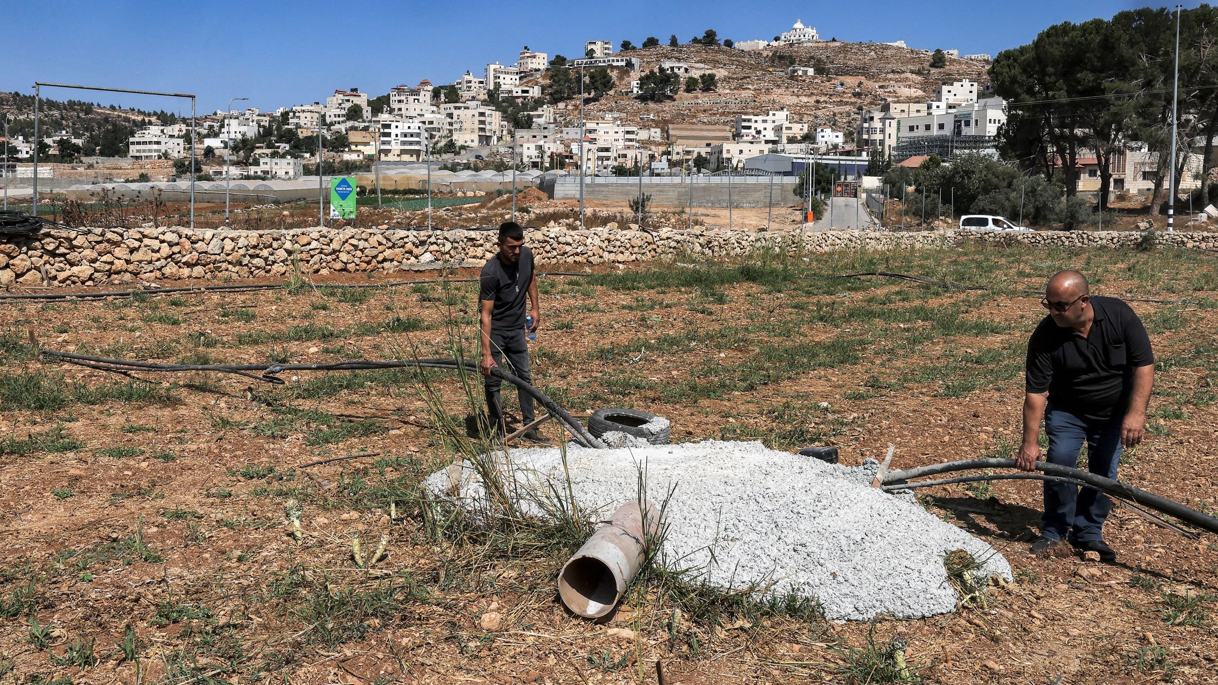 در این تصویر، دو مرد در حال کار بر روی یک زمین کشاورزی هستند. آنها مشغول بررسی یا نصب لولههایی هستند که به یک سازه بتنی متصل شده است. درختان و گیاهان در اطراف وجود دارند و در پسزمینه، ساختمانی و تپههایی دیده میشوند که حاکی از یک منطقه مسکونی است. آسمان آبی و صاف به نظر میرسد.