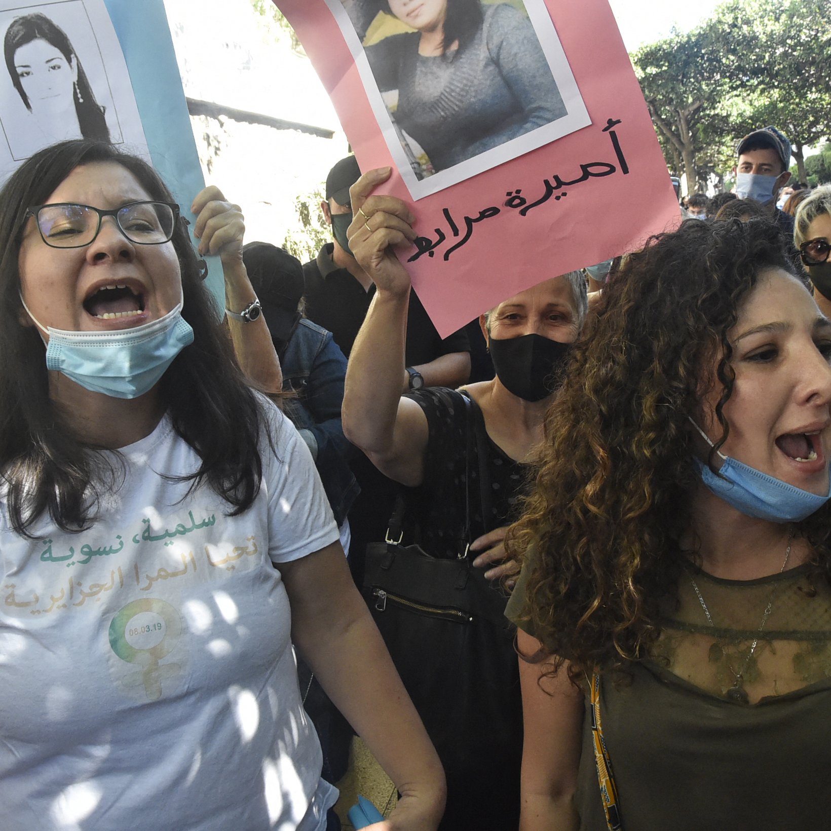 L'image montre un groupe de manifestants lors d'une manifestation. Plusieurs personnes tiennent des pancartes avec des photographies et des slogans. Les participants semblent exprimés des émotions fortes, comme la colère ou la détermination, en scandant des slogans. Certaines personnes portent des masques de protection, tandis que l'environnement semble urbain avec des arbres et des bâtiments en arrière-plan.
