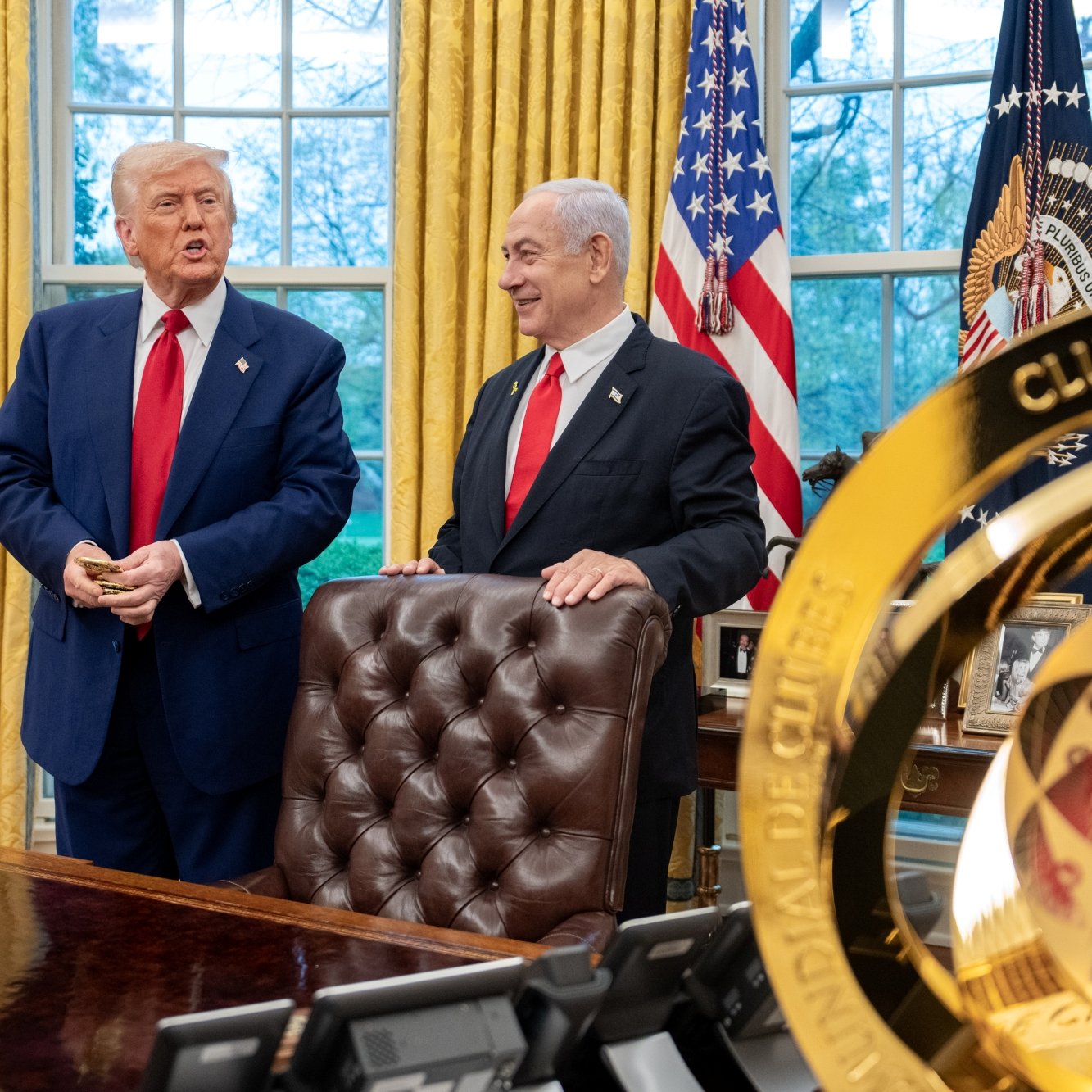 Deux hommes souriants devant un bureau, drapeaux en arrière-plan, avec un trophée doré au premier plan.