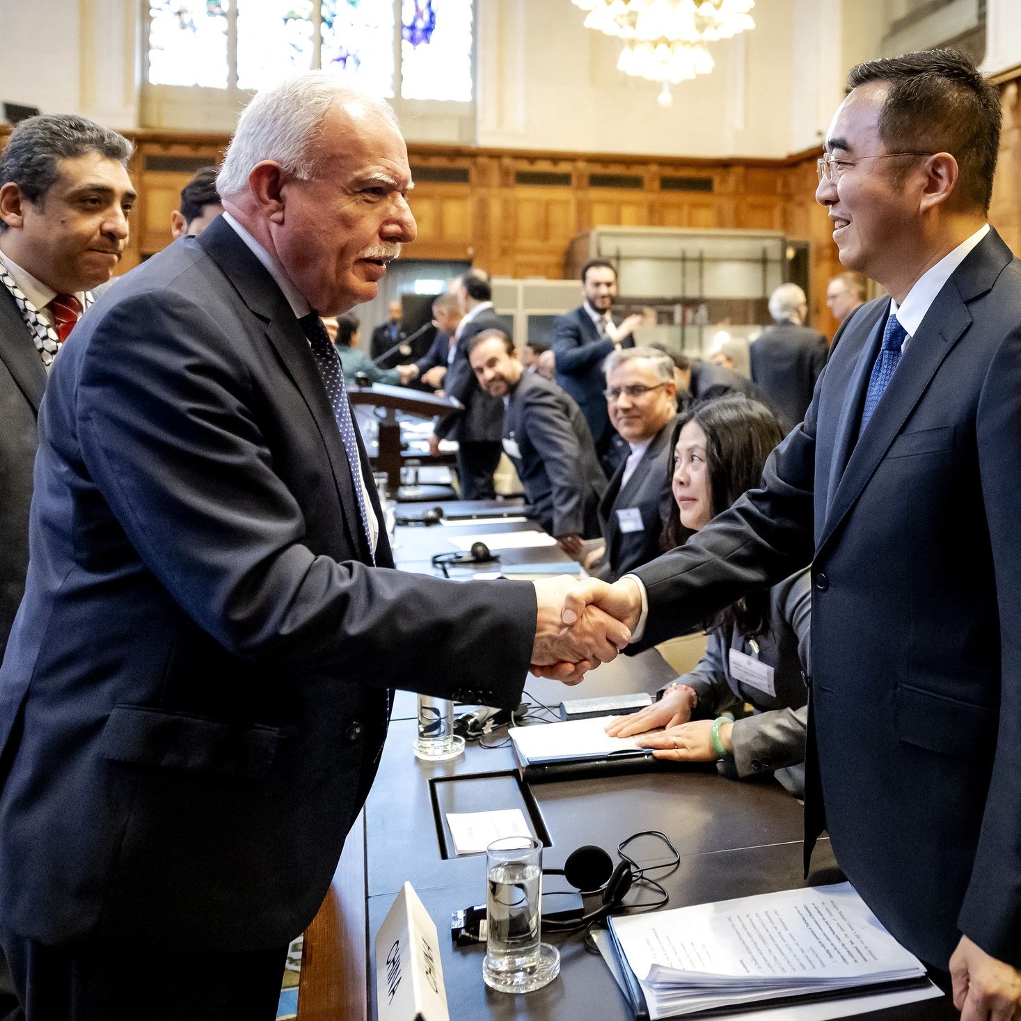 The image depicts a formal gathering, likely a conference or meeting, with several individuals present at a large table. In the foreground, two men are engaged in a handshake, symbolizing a greeting or agreement. One of the men appears to be in a dark suit, while the other is also in formal attire. The background features wooden paneling and other participants seated at the table, some of whom are interacting with each other. The setting suggests a professional and diplomatic atmosphere.