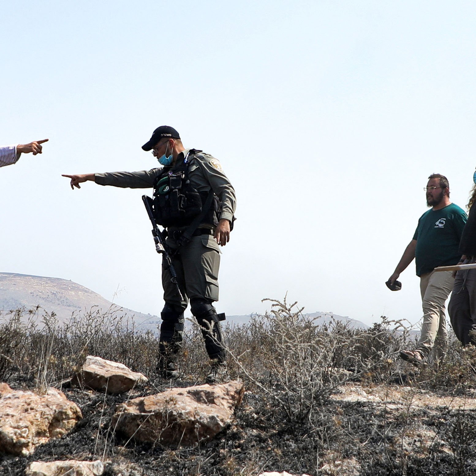 L'image montre une scène de tension entre un homme habillé dans un style traditionnel, tenant une sorte de bâton ou d'objet, qui semble pointer du doigt. Un policier ou un soldat, portant un uniforme et un gilet, est en position d'affrontement avec lui, pointant également vers l'homme. En arrière-plan, plusieurs autres personnes, visiblement des observateurs ou des participants à la scène, se tiennent. Le paysage environnant semble aride, avec des rochers. L'atmosphère est chargée et indique une situation potentiellement conflictuelle.