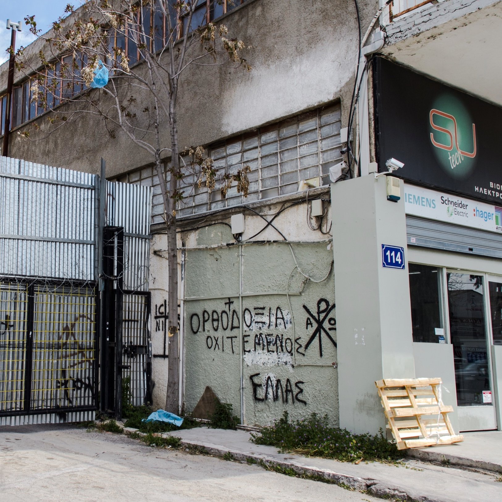 L'image montre un environnement urbain avec un bâtiment en arrière-plan et un mur métallique en façade, où l'on peut voir des graffitis. Un drapeau grec est visible, ainsi qu'un espace de stationnement ou de stockage à côté d'une entrée. Le sol semble en béton, et quelques déchets sont éparpillés sur le sol. Les graffitis sur le mur comprennent des inscriptions en grec, ce qui suggère un contexte local ou culturel. L'atmosphère paraît un peu désordonnée, typique des zones industrielles ou abandonnées.