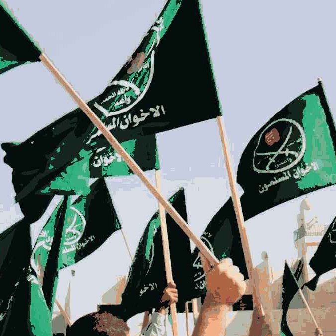 L'image montre un groupe de personnes levant des drapeaux verts. Les drapeaux semblent porter des inscriptions en arabe ainsi que des symboles, indiquant une affiliation à un mouvement ou une organisation spécifique. L'ambiance semble dynamique et engagée, suggérant un rassemblement ou une manifestation. L'arrière-plan indique probablement un espace public ou un lieu symbolique.